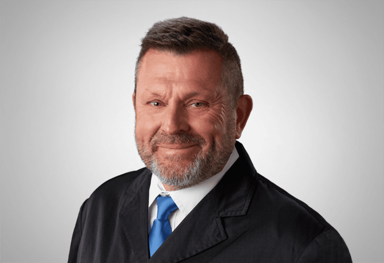A man in a white shirt, black suit jacket and blue tie is smiling, with a grey background behind him.