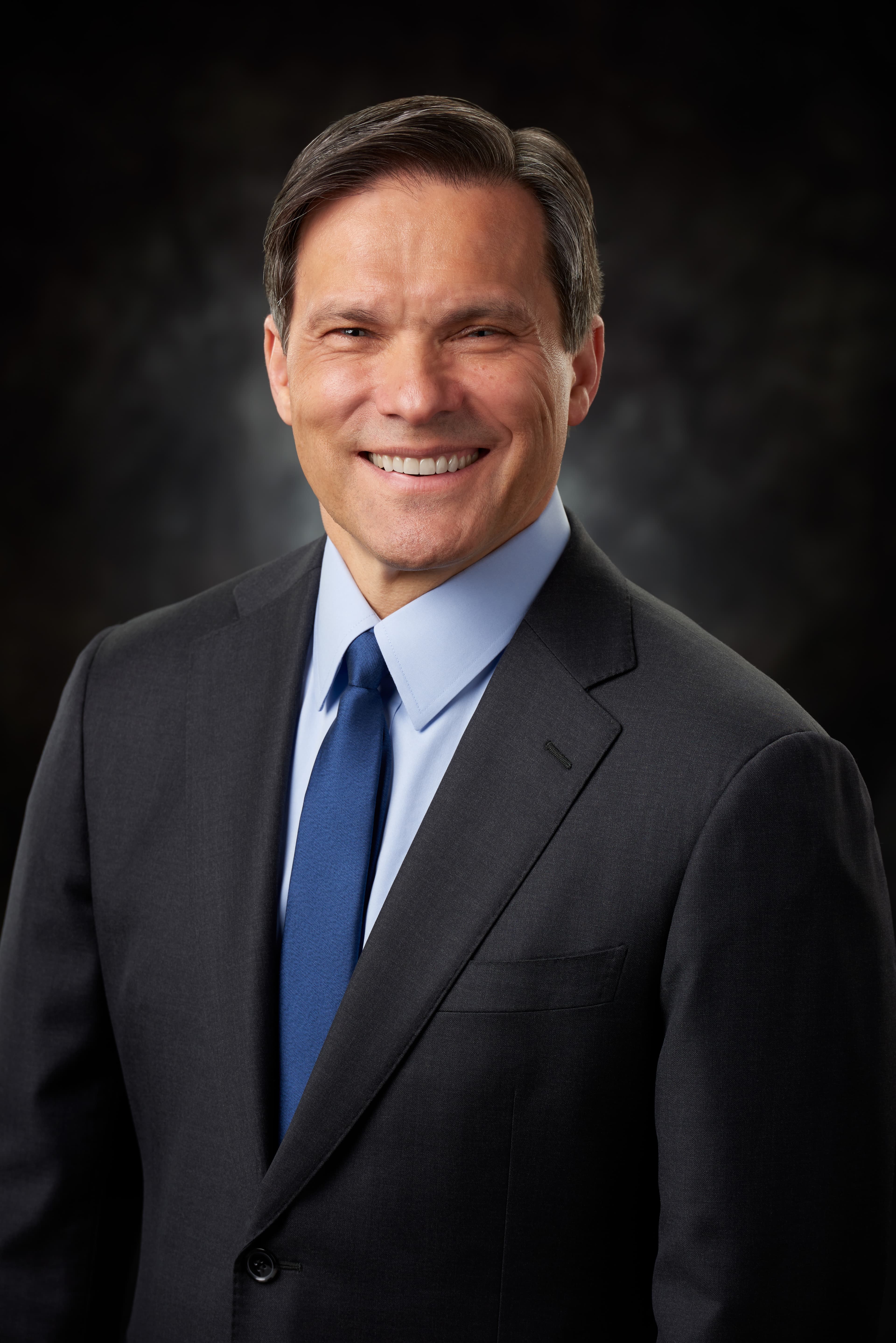 A man wearing a suit and blue tie smiles in front of a black background.