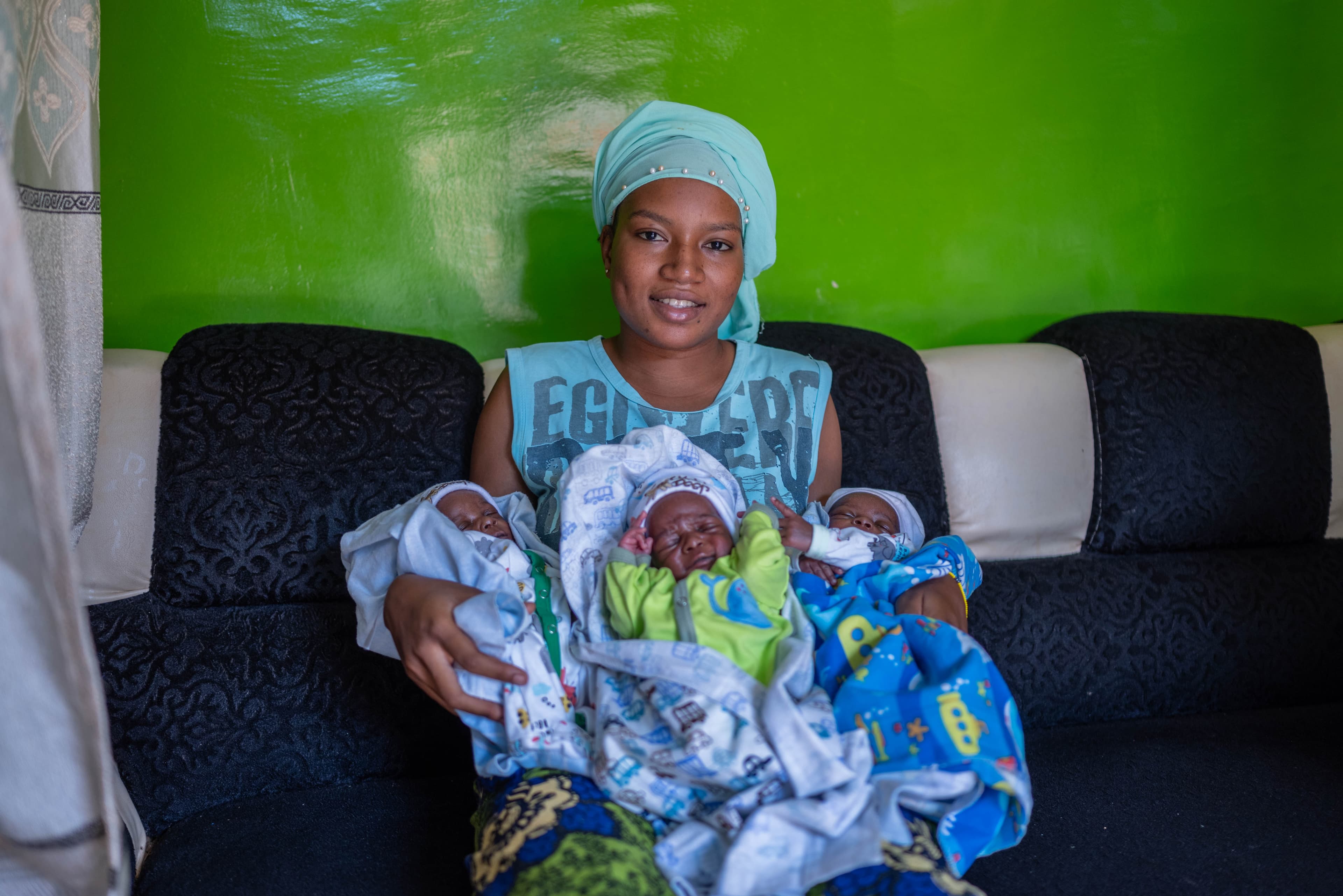 Young mother wearing a light blue head scarf and shirt sits with her triplets, wrapped in blankets.