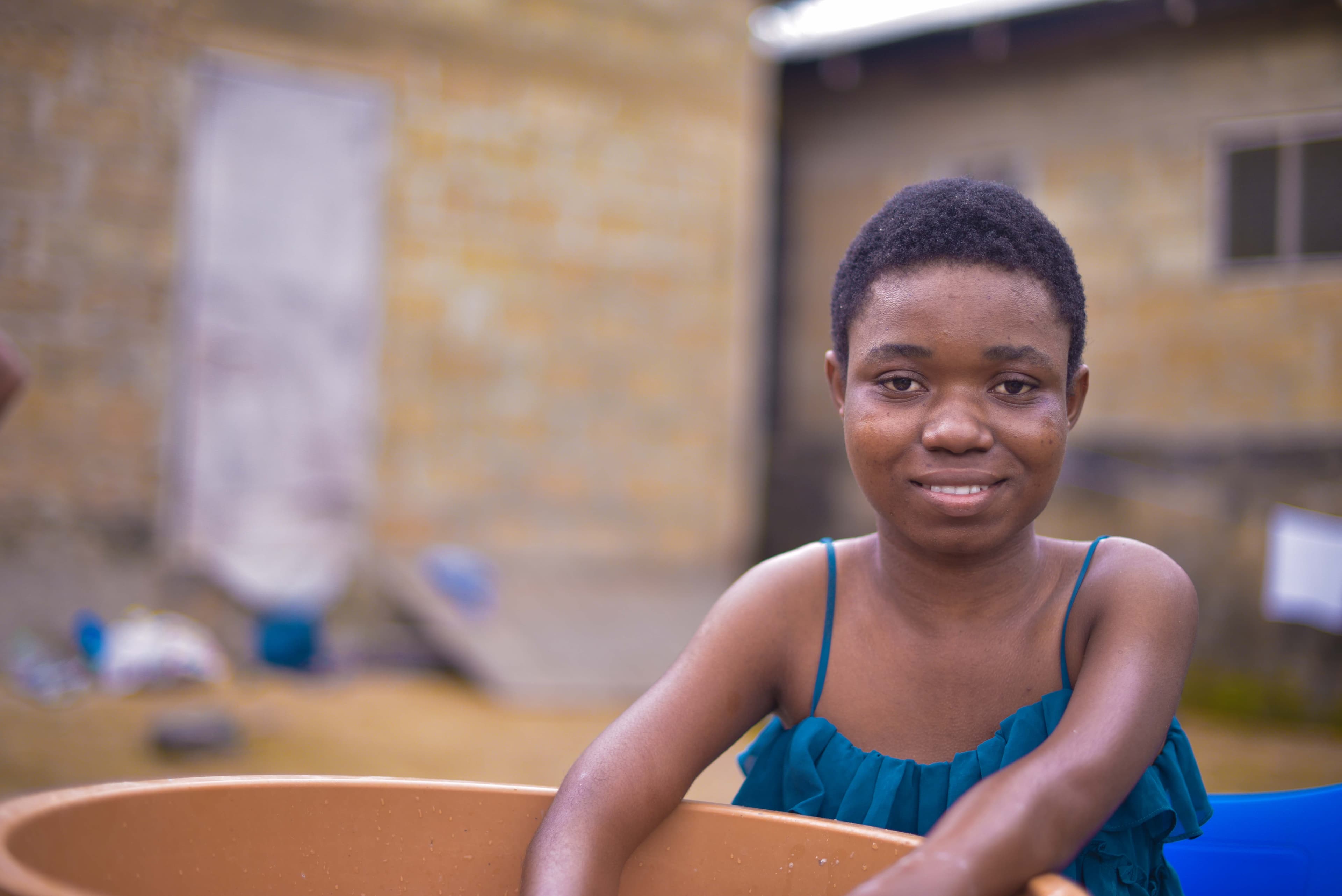A teen African girl wearing a blue shirt smiles for the camera with her hands in a bucket.