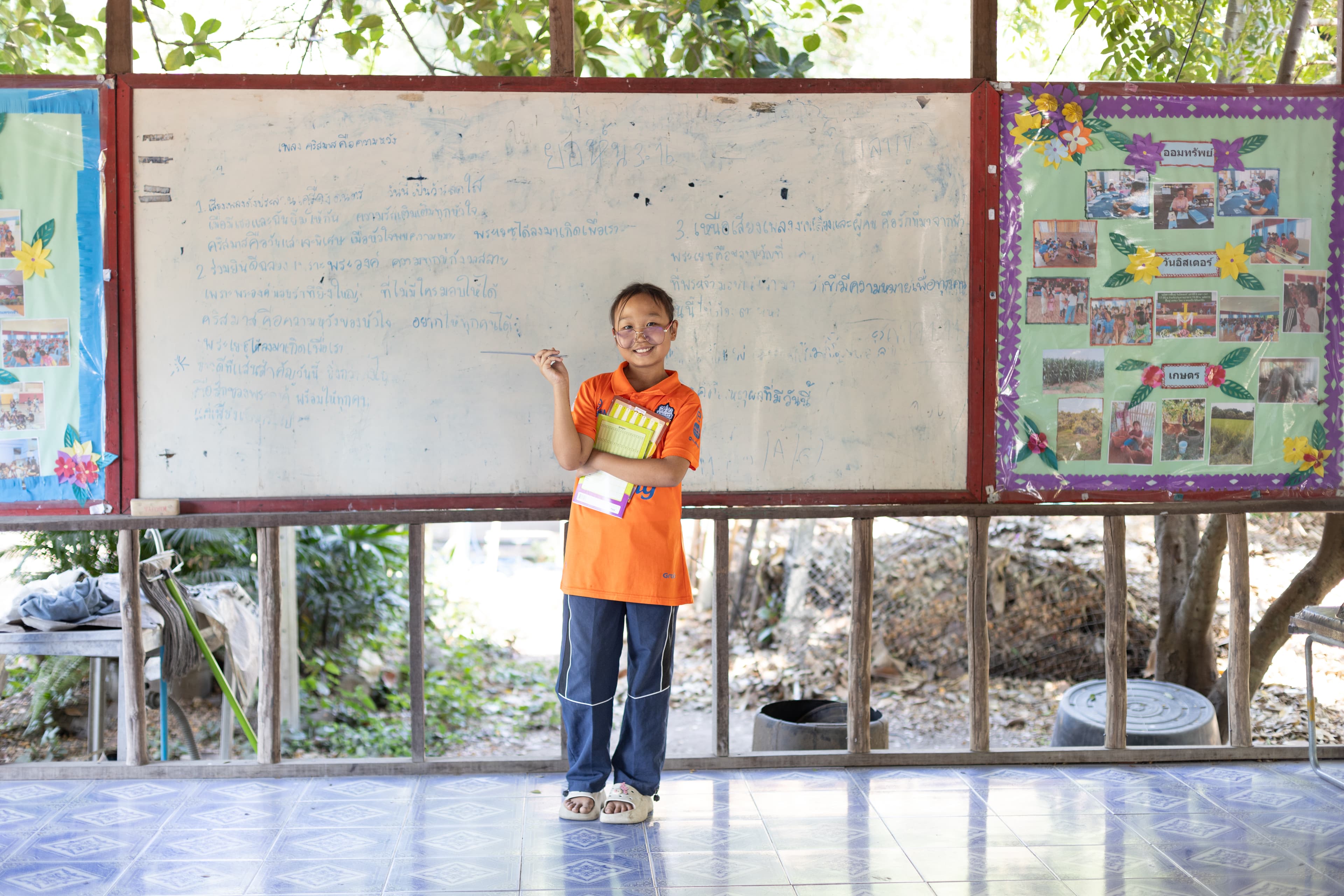 Young, Thai girl wearing an orange shirt and blue pants stands in front of a white board, pretending to be a teacher.