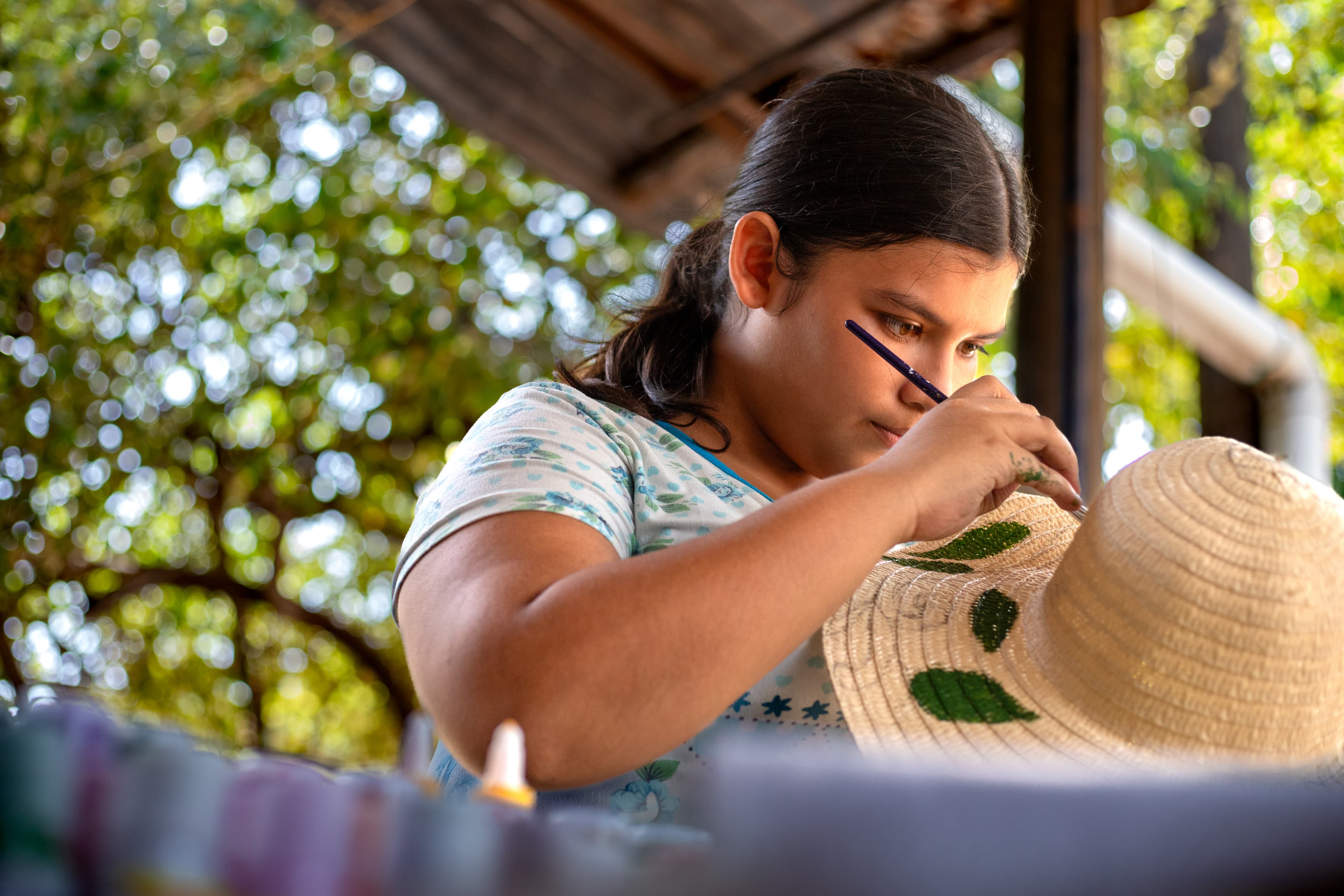 A teenage girl wearing a blue and white shirt paints green leaves on a straw hat with a black paintbrush.
