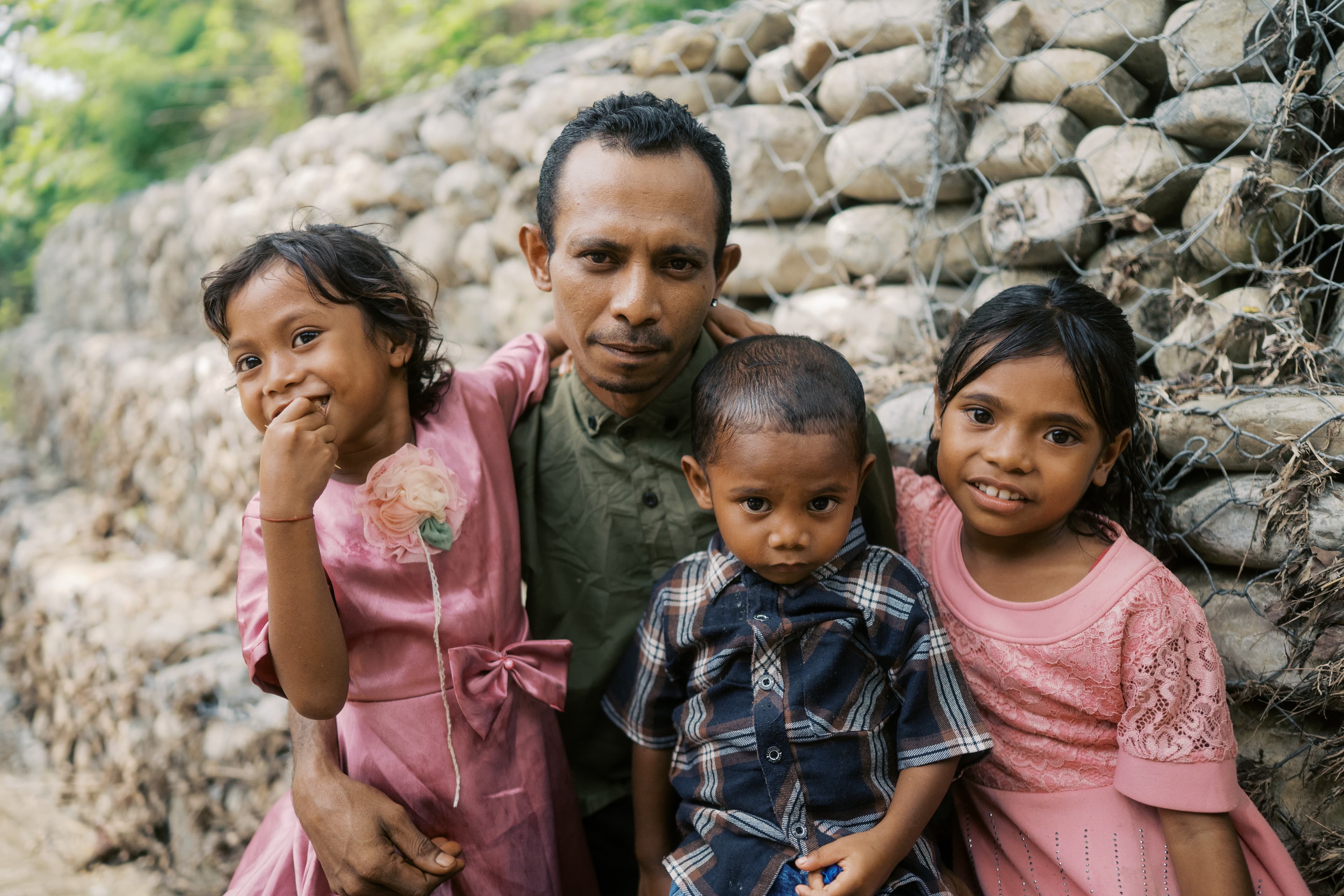 A father sits with his three children in front of a rock wall covered in a metal fence.