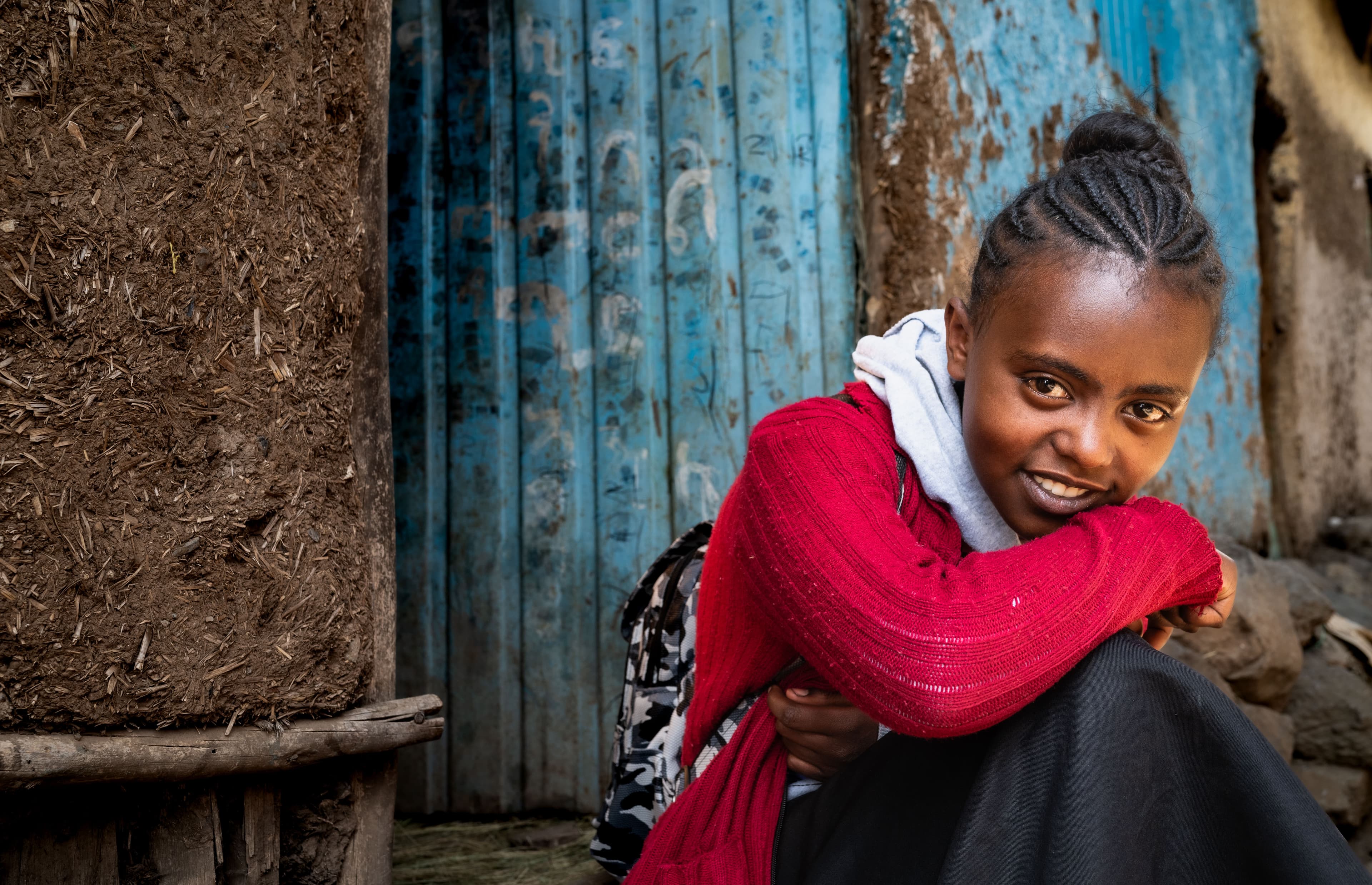 A young girl wearing a red jacket sits in front of a wall made of dirt, wood and blue paint.