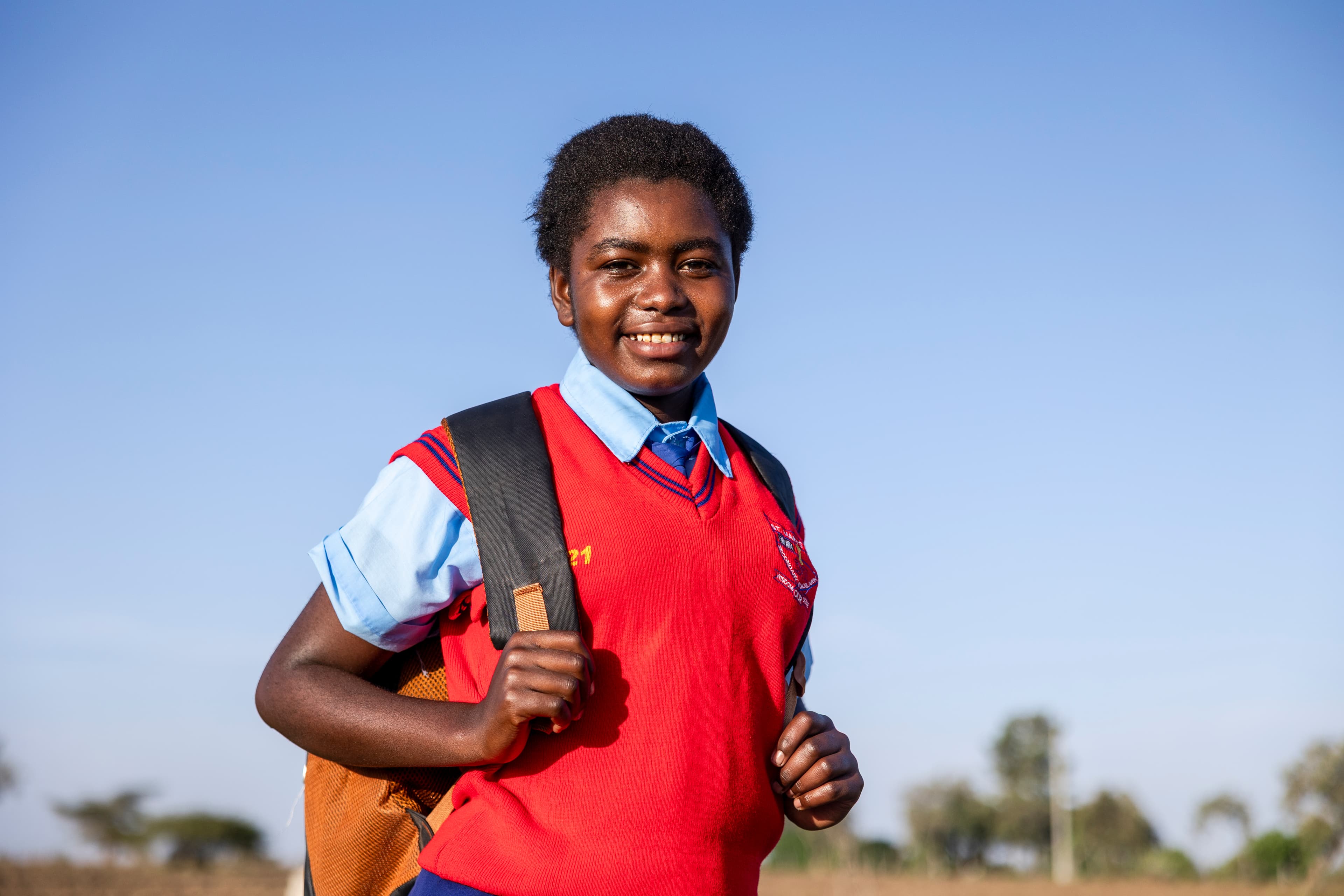 A young girl is standing in a field with her hands holding her backpack.