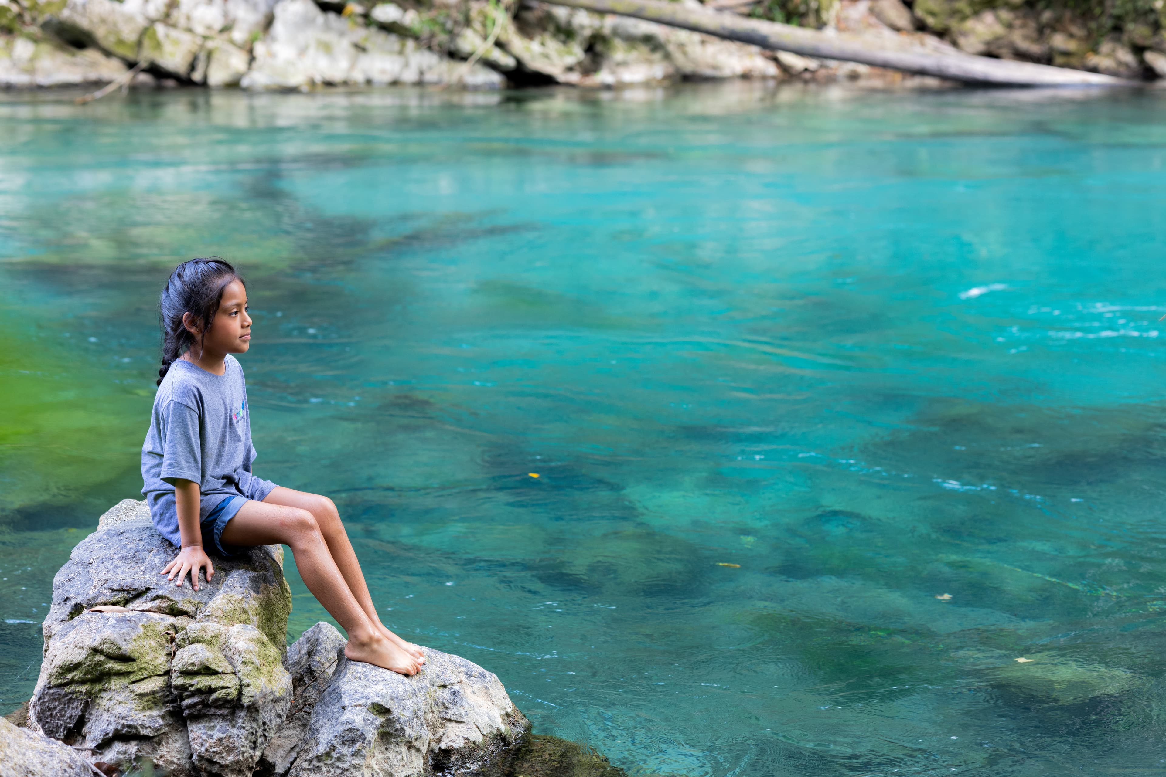 A young girl sits on a rock in front of a bright blue lake.