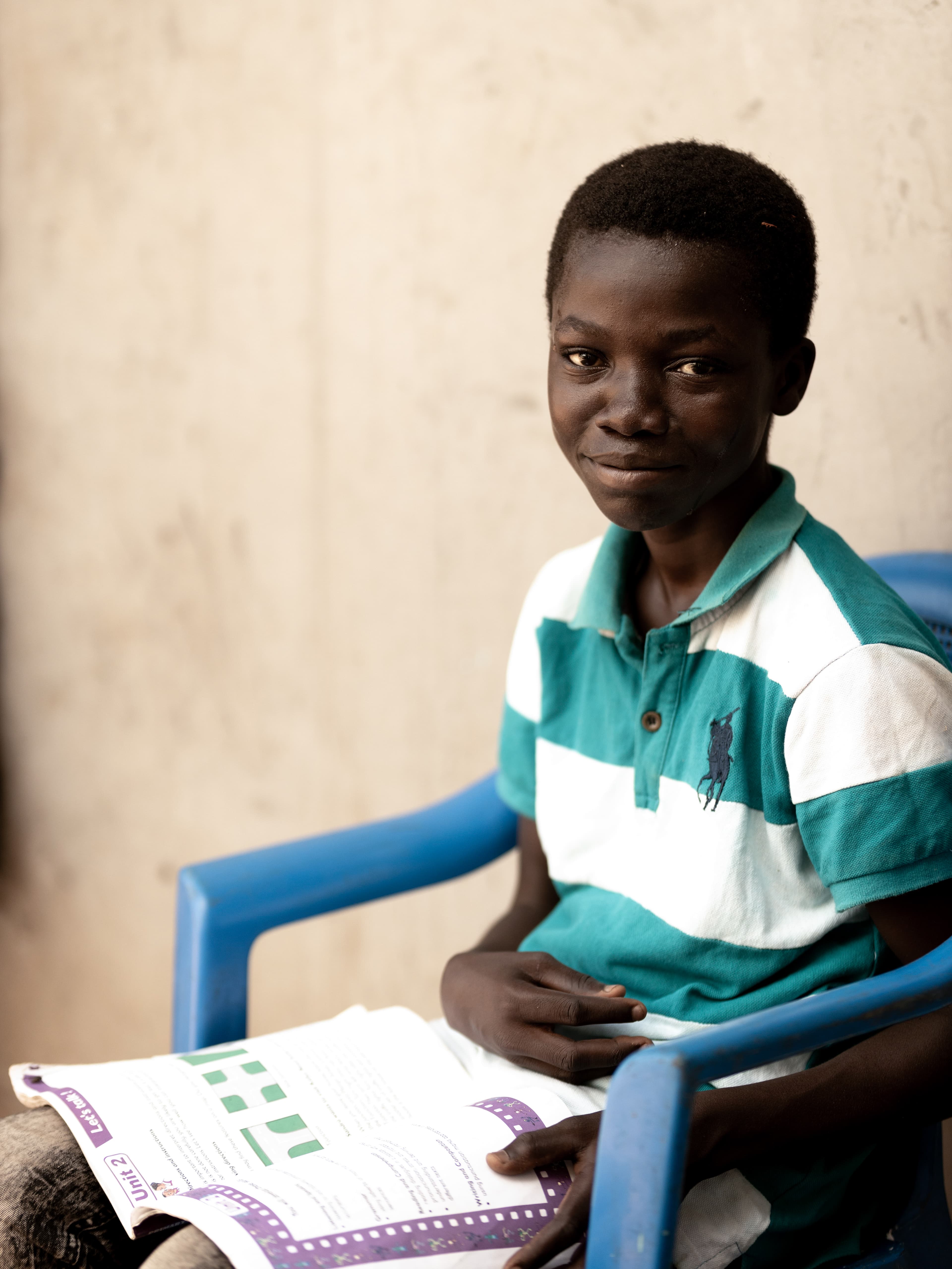 A teen African boy wearing a striped shirt sits in a chair and smiles for the camera.