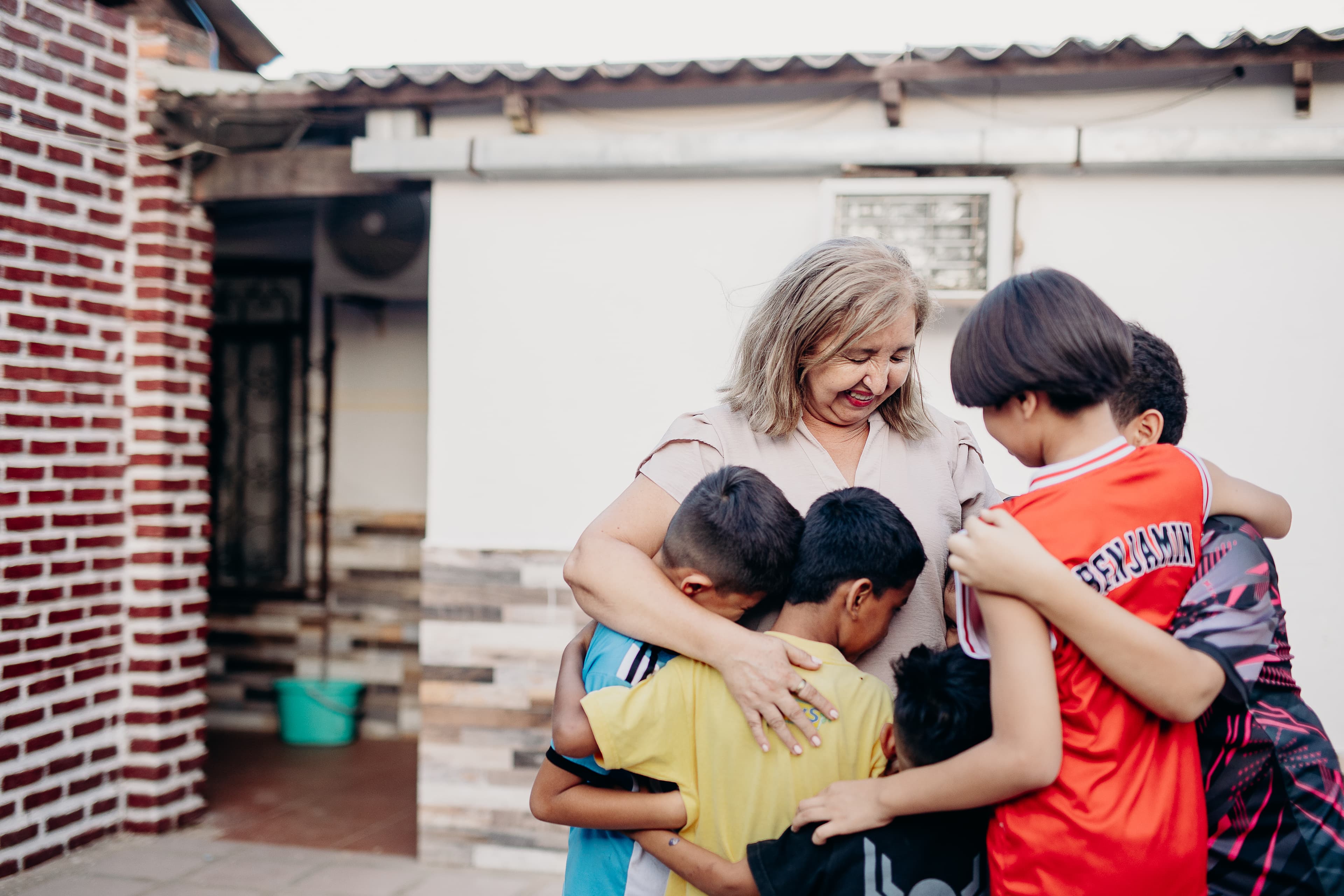 Woman smiles while embracing four young boys in front of a brick building.