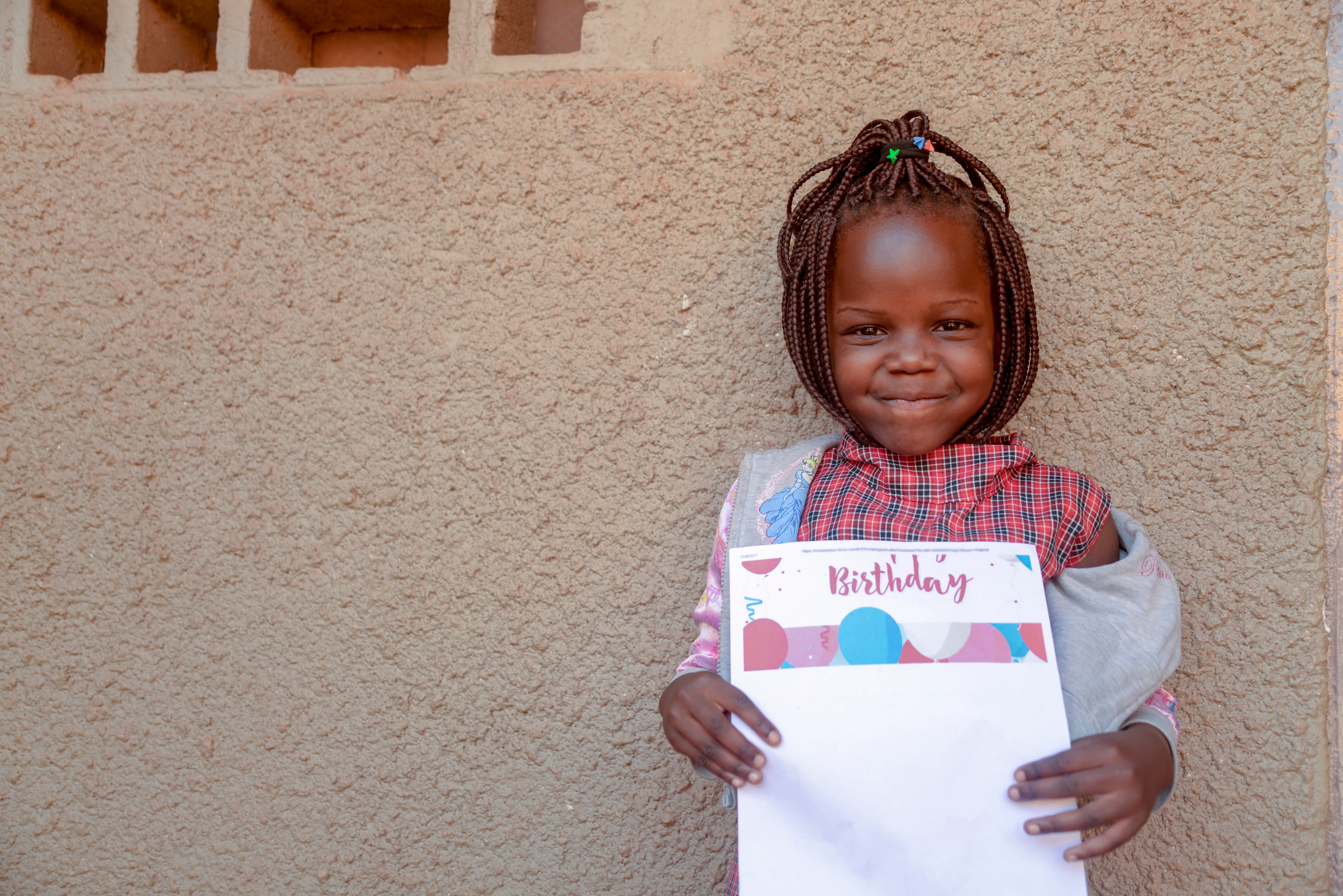 A young African girl stands in front of a concrete wall while holding a sponsor letter and smiling.