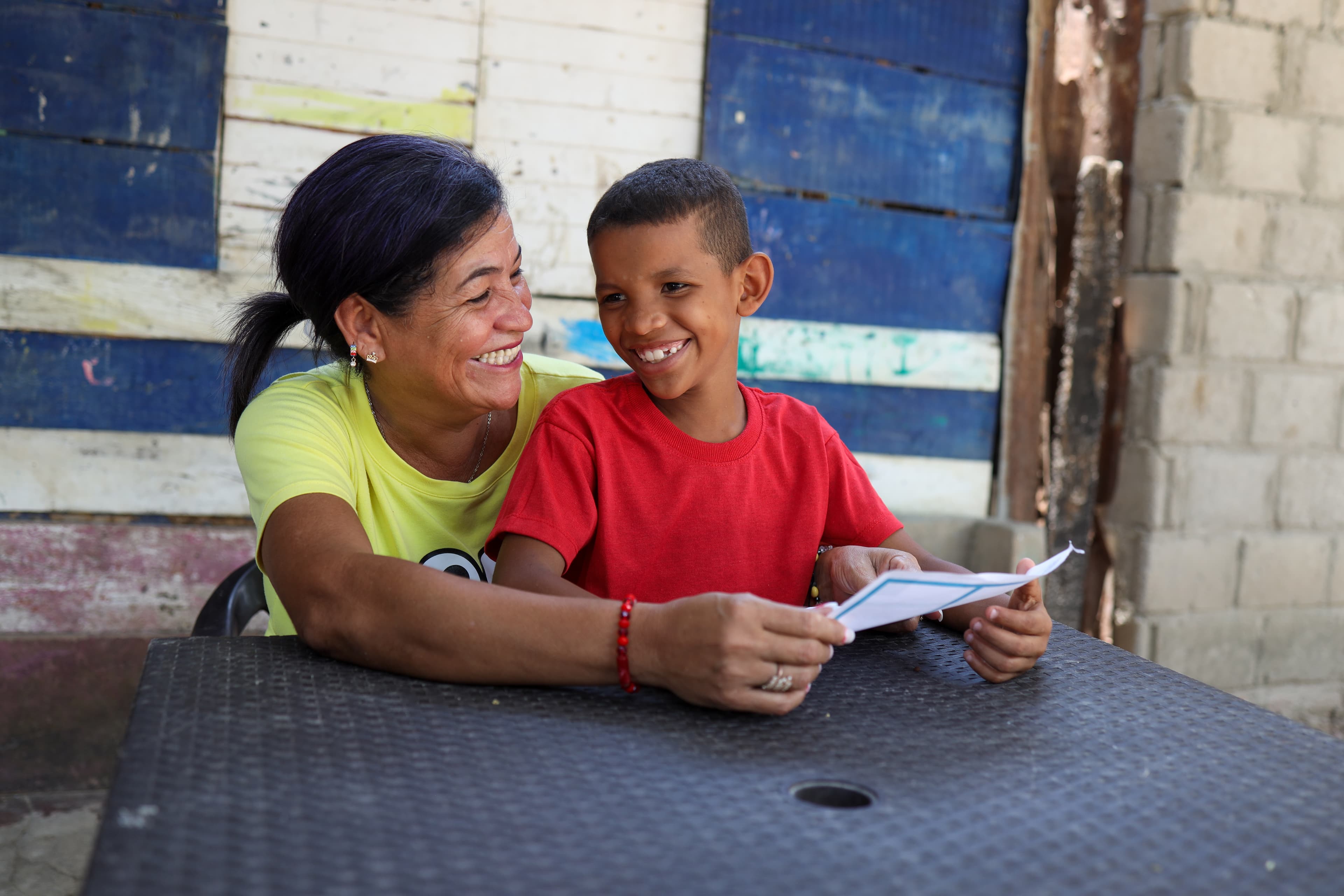 Colombian boy wearing a red shirt smiles while sitting at a table with his mother.