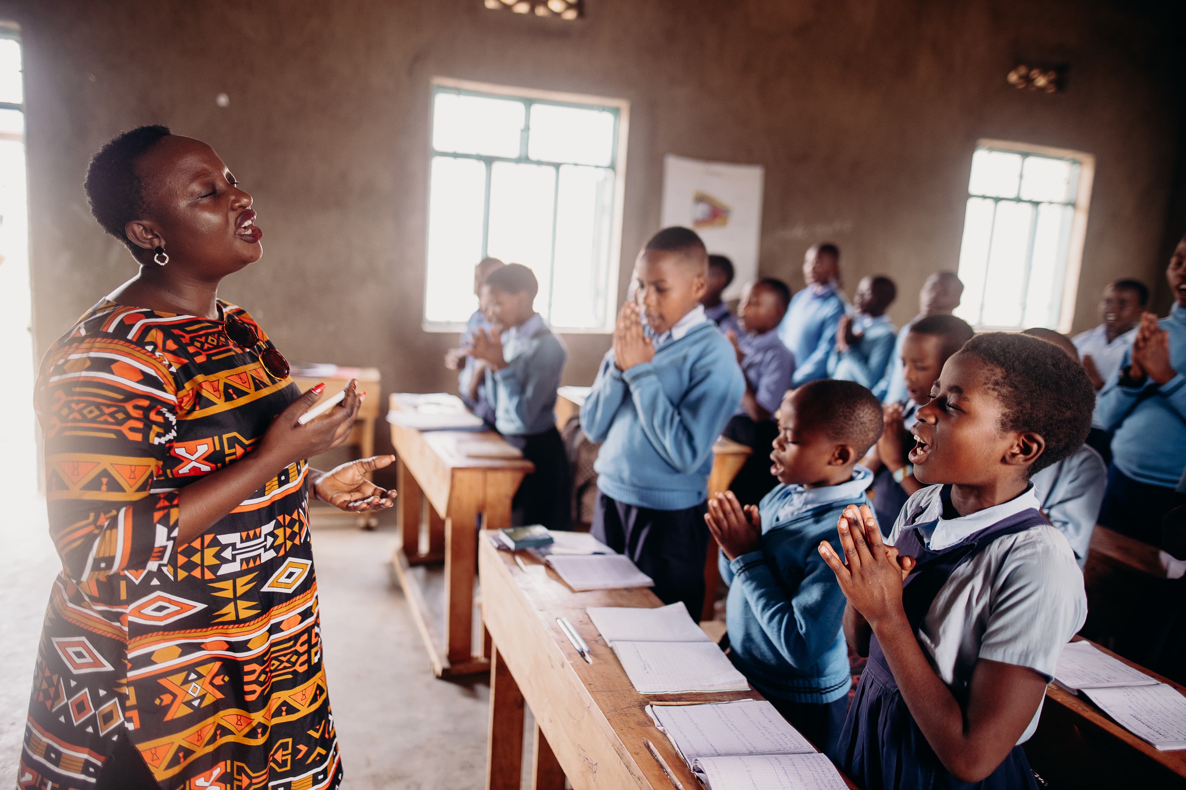 Woman wearing a patterned shirt and black pants leads a classroom full of Rwandan students in prayer.