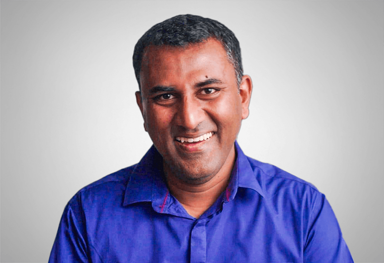 A man in a blue button up shirt smiles, with a grey background behind him.