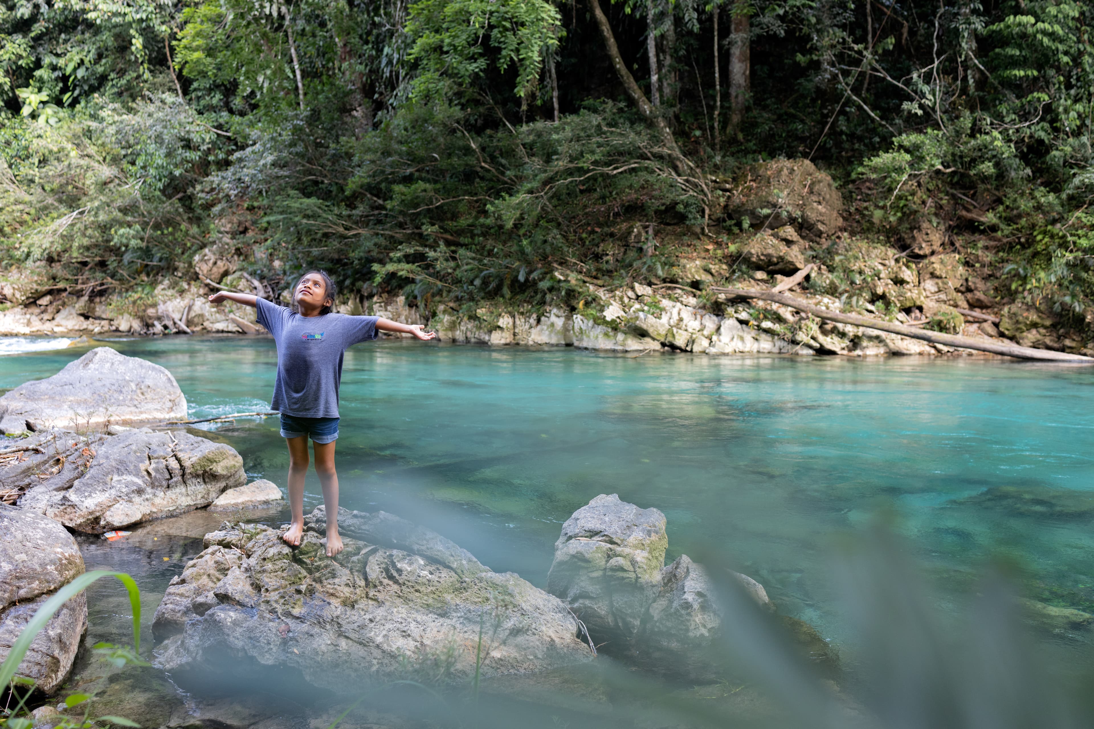 A young girl stands on a rock in a lake with her arms outstretched wide.