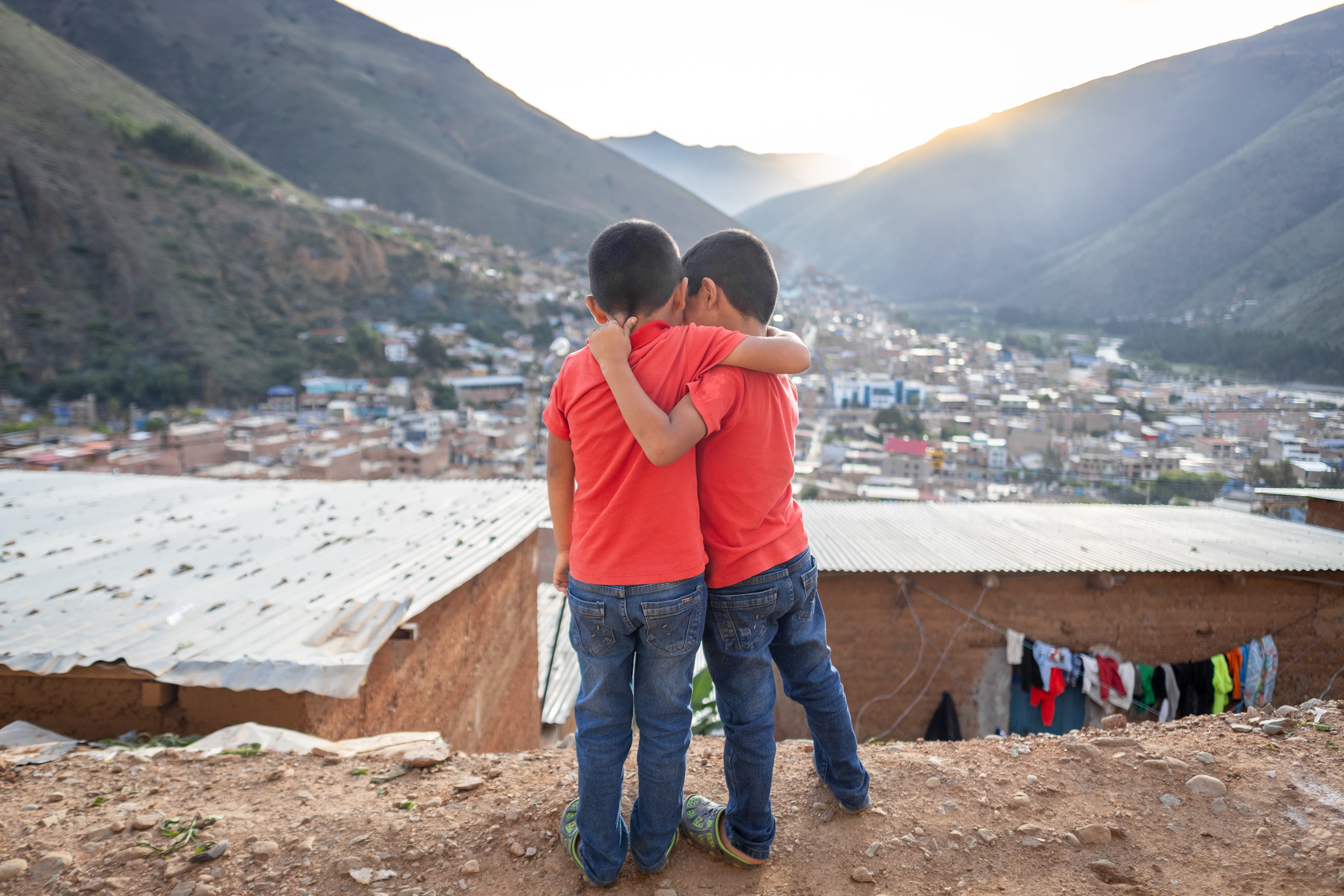 Two young boys wearing red shirts embrace in front of a sunset behind mountains.