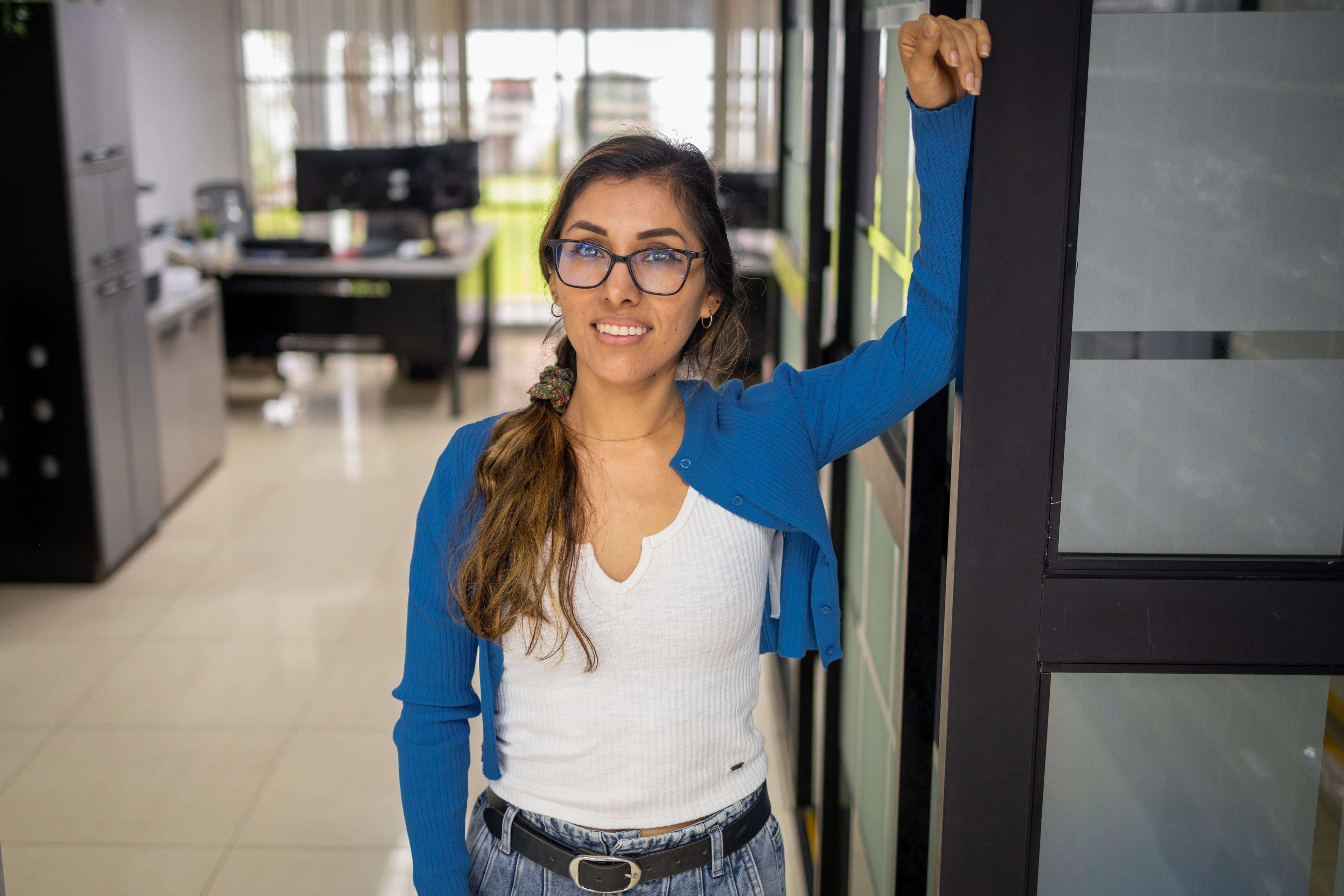 A young woman stands against a door with her arm lifted as she smiles at the camera.