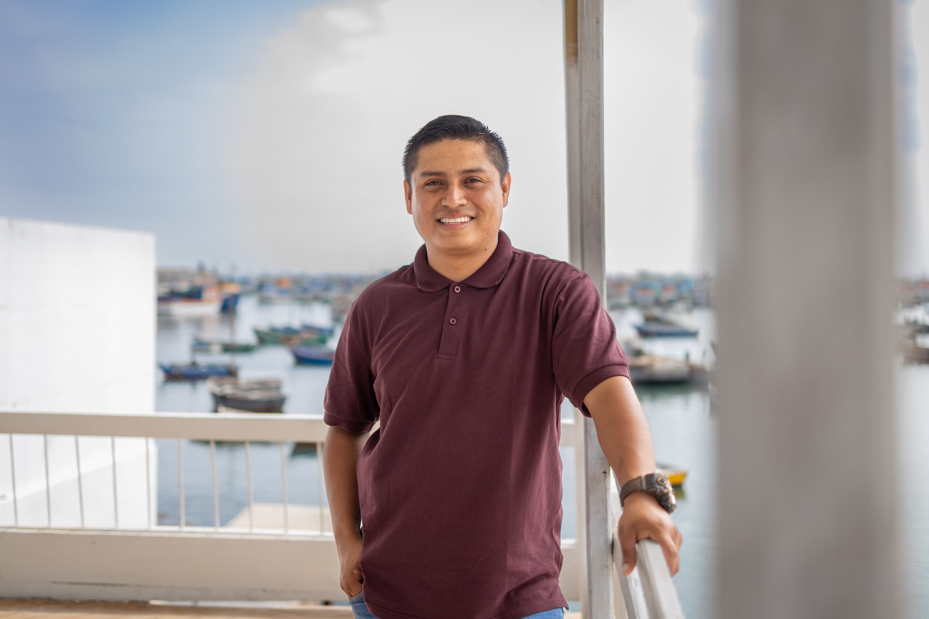 A man stands with his hand on a railing overlooking the water as he smiles at the camera.