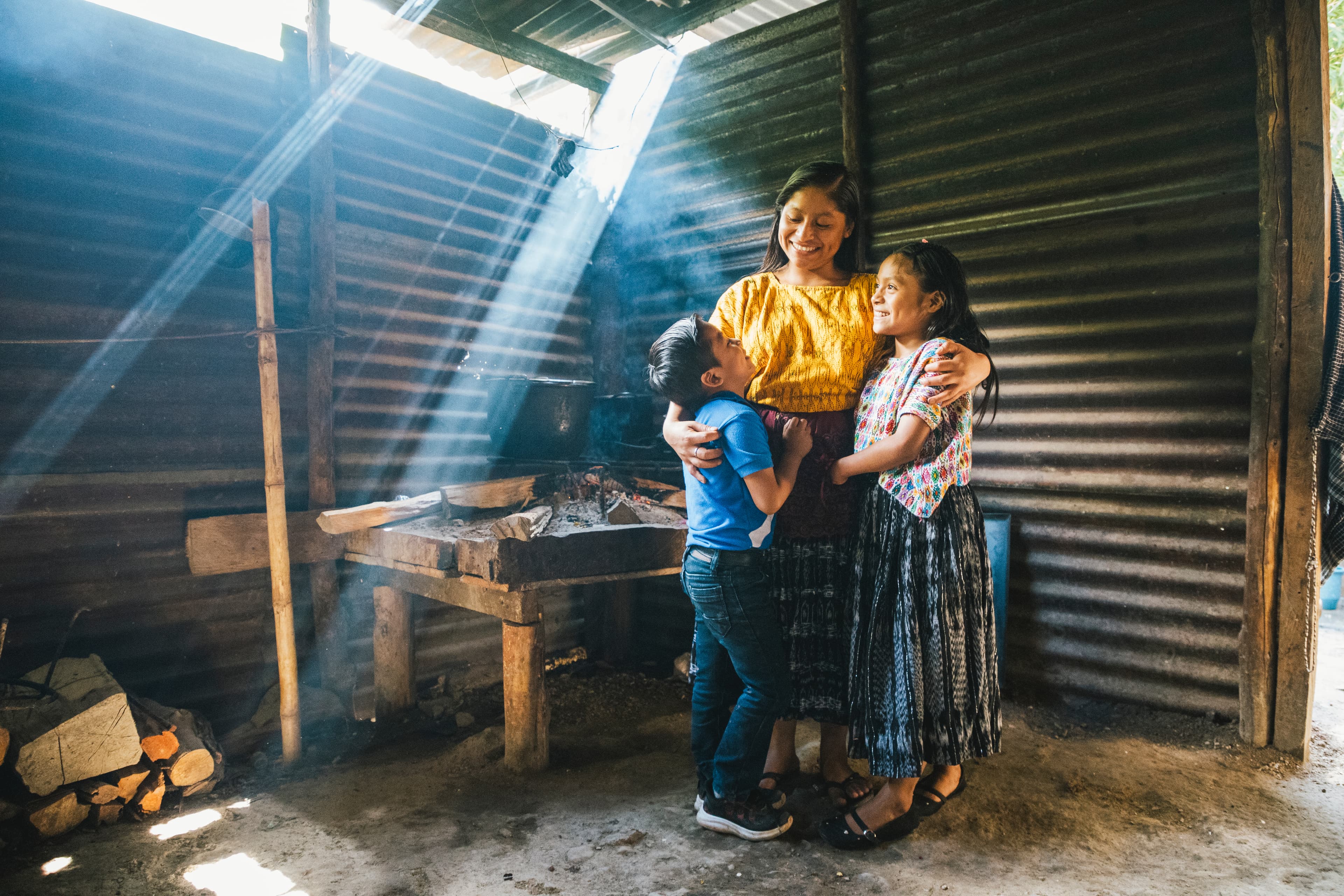 A mom embraces her two children in front of a tin wall as the sun streams in.