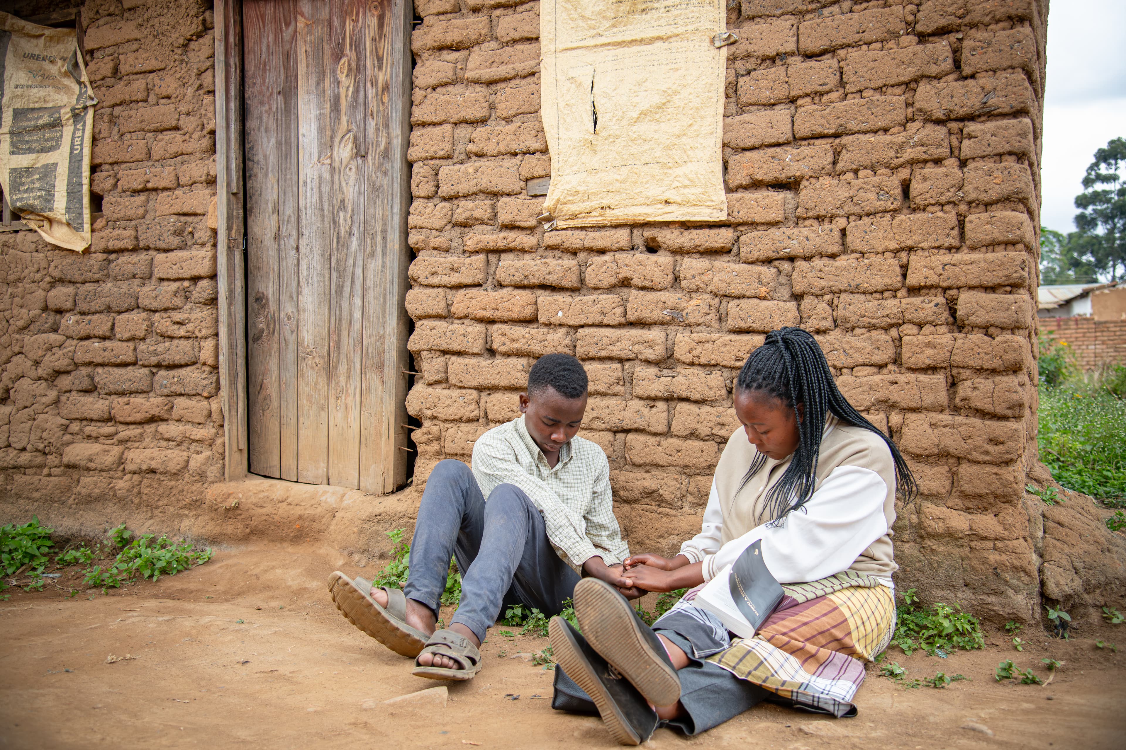 A young boy is sitting on the ground outside his house holding hands and praying with his mentor.