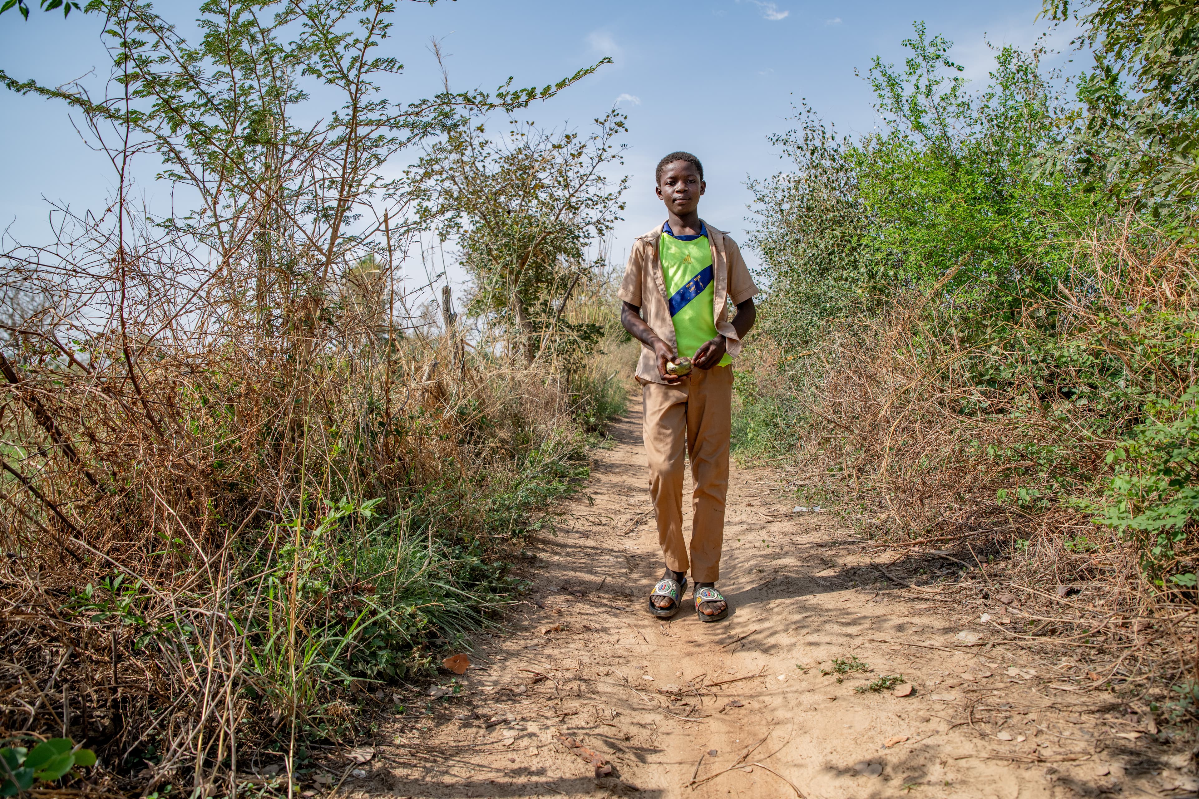 A young boy walks down a dirt path surrounded by trees and grass.