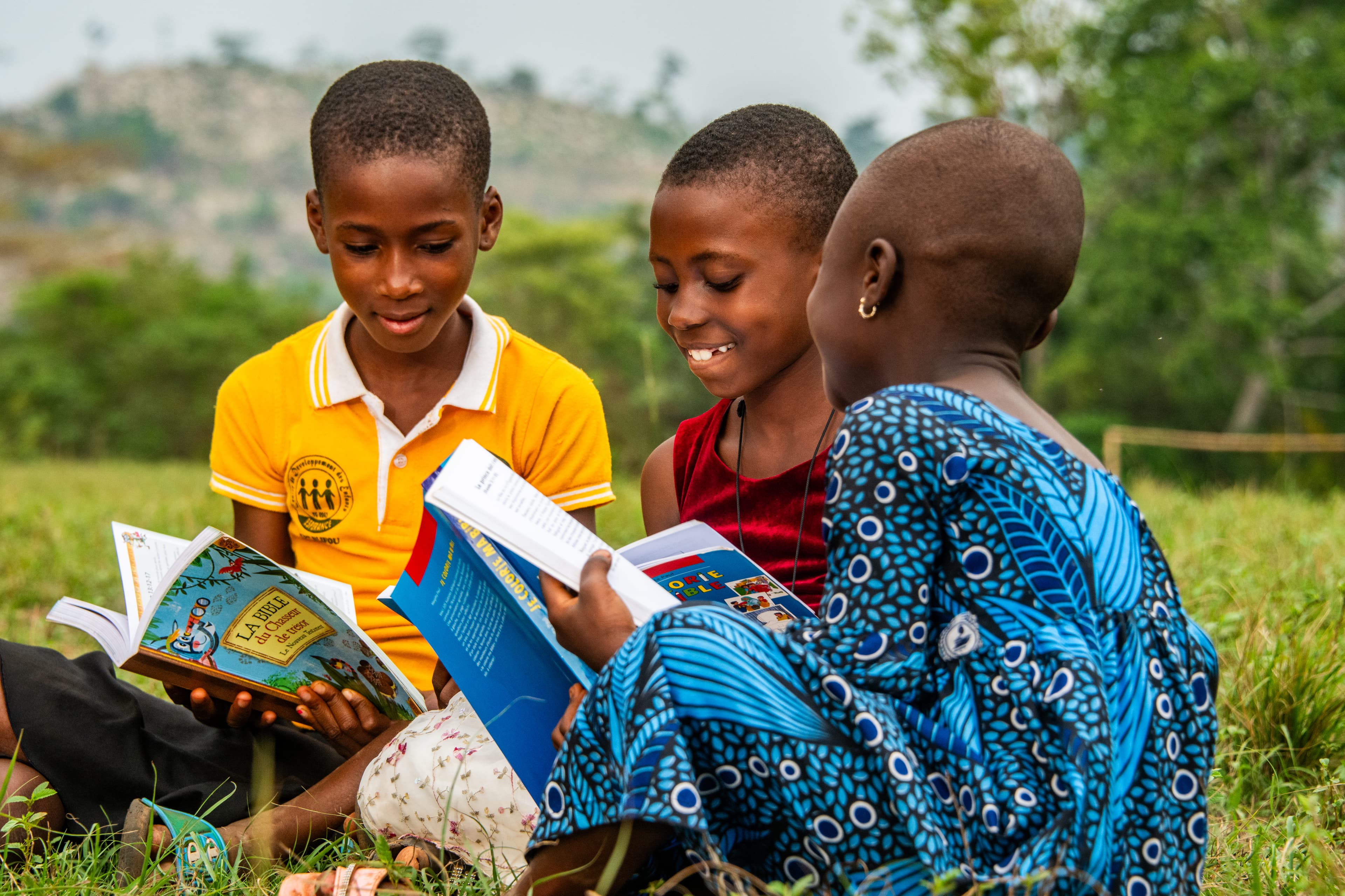 Three young African girls sit in the grass while reading their Bibles and smiling.