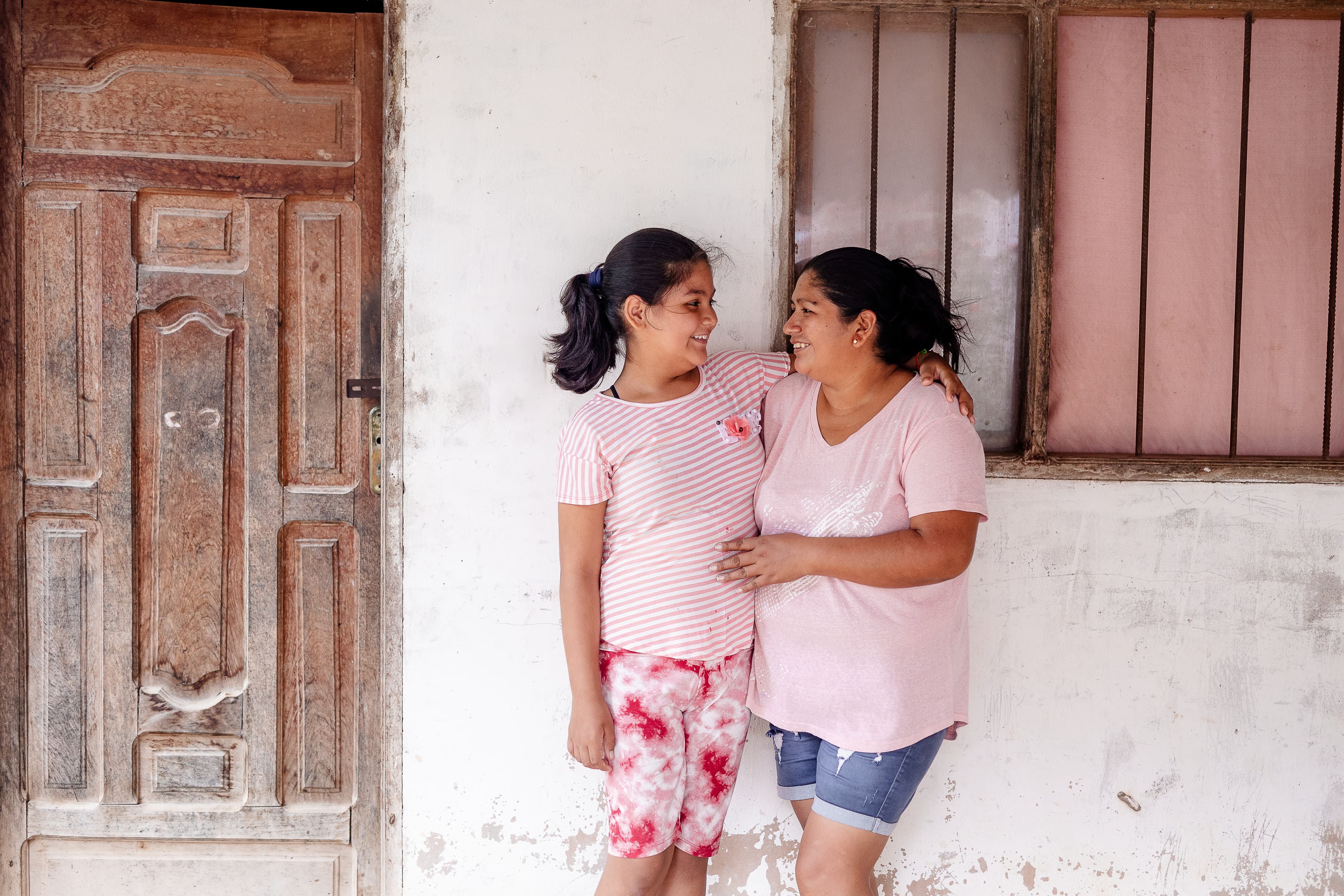 A young girl and her mom embrace and smile at each other outside their home.