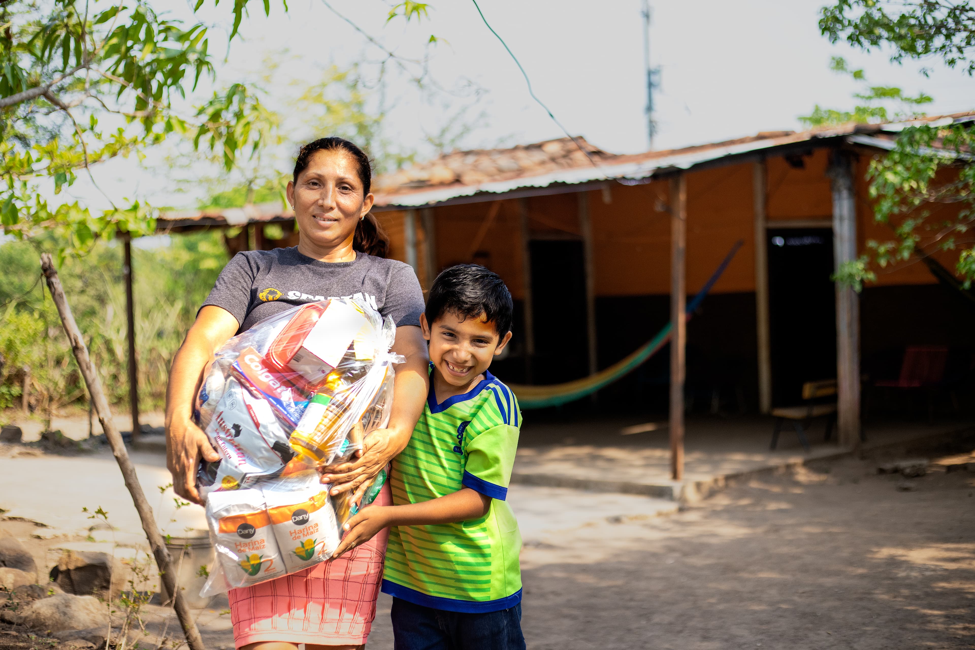 Smiling mother holds bag of food while standing with son outside home.