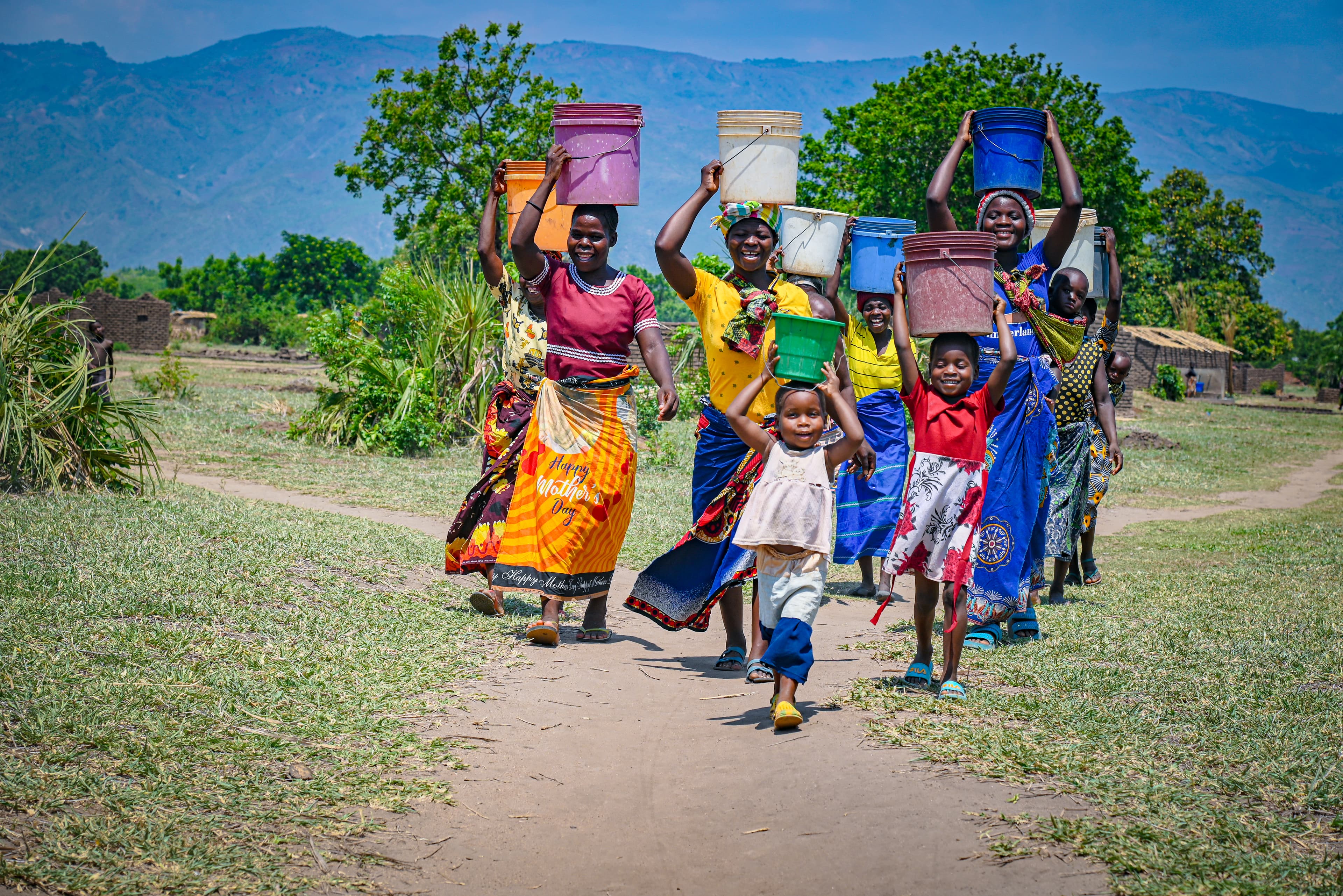 A child leads a group of women and children walking along a dirt path. They are all carrying buckets of water on their heads. The buckets are orange, purple, white, green, and blue. There are trees, small brick homes, and mountains in the background.