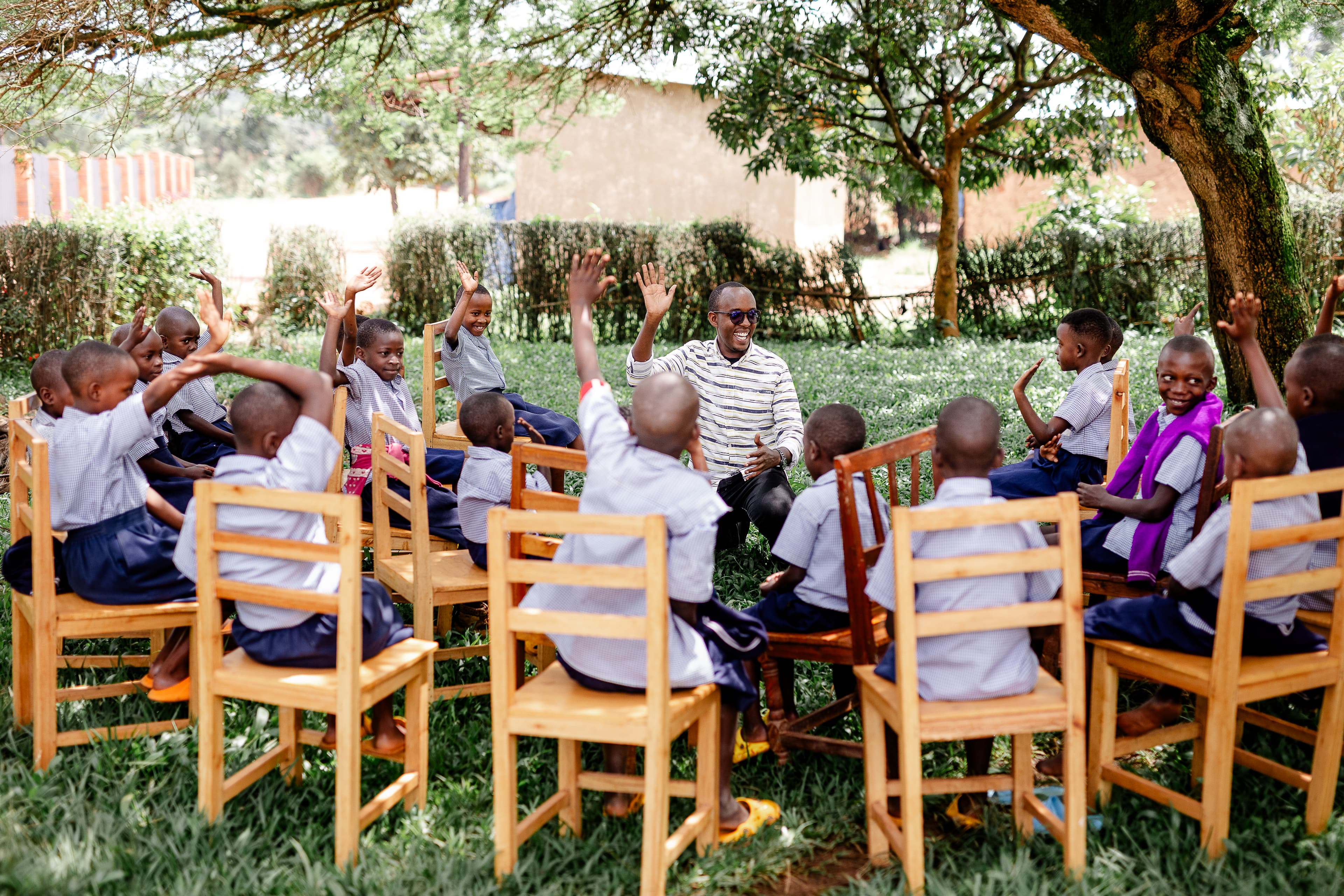 An African man sits in the middle of a group of children in chairs as they all raise their hands.