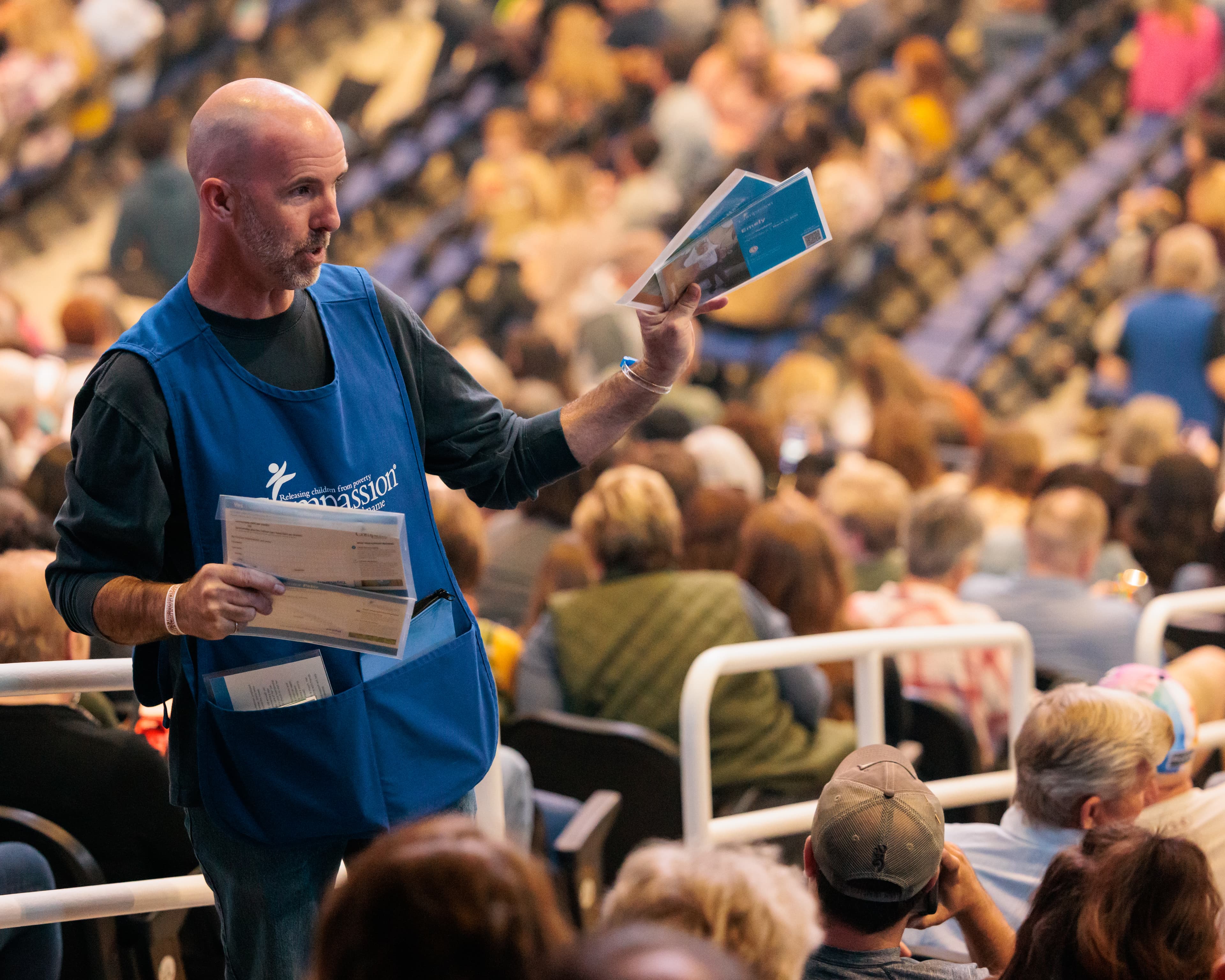 A man in a blue volunteer apron holds child sponsor cards in his hands, while walking in a large auditorium full of people.
