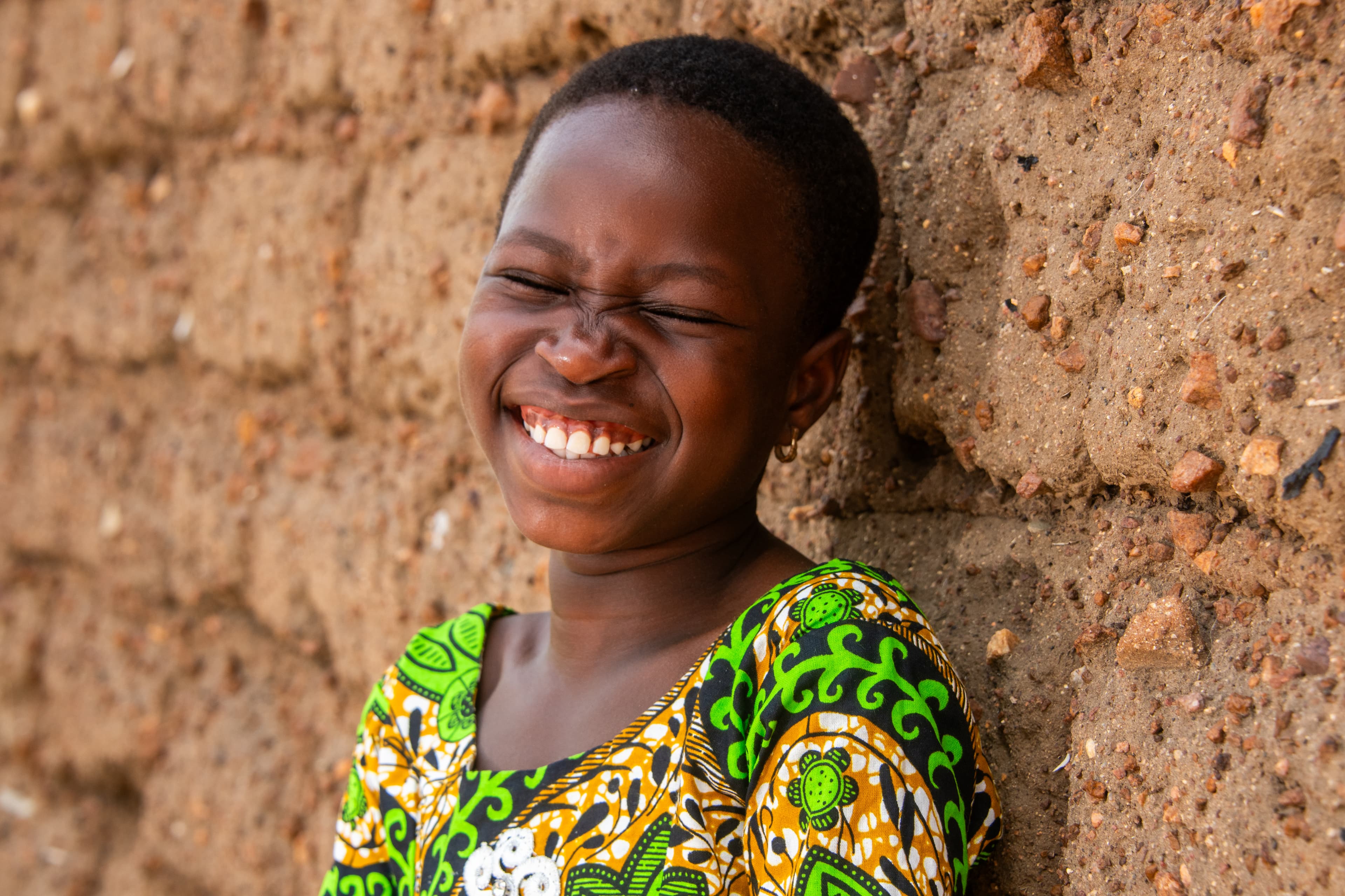 Young African girl smiles with her eyes closed in front of a clay wall.