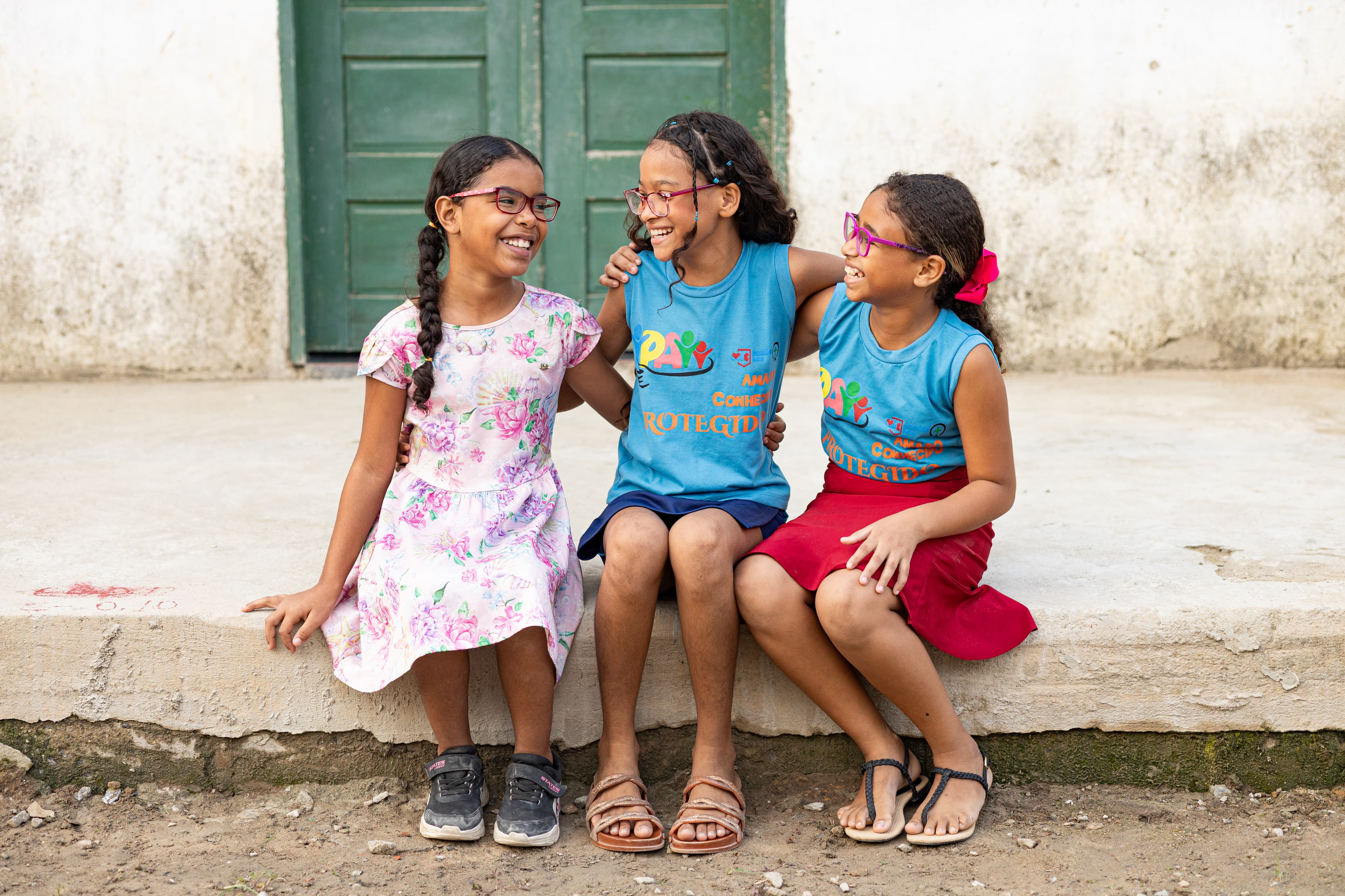 Three young girls are sitting on a sidewalk smiling with their arms around one another.