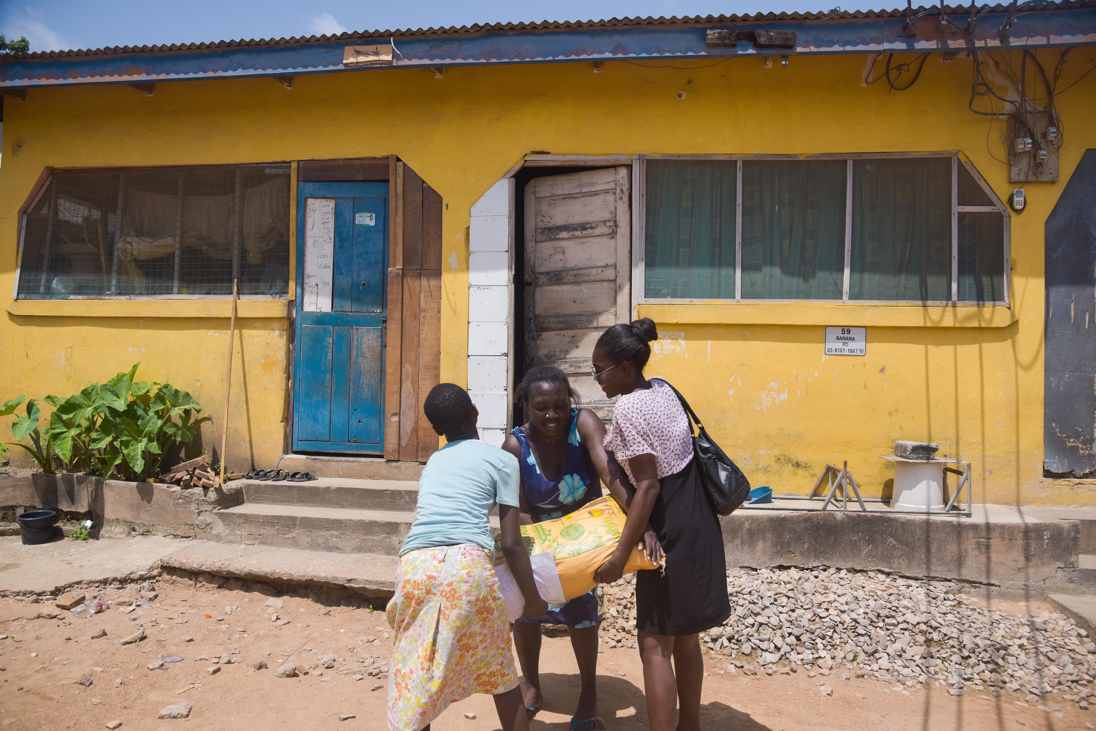 Two African women give a bag of food to another African woman in front of a bright yellow house.