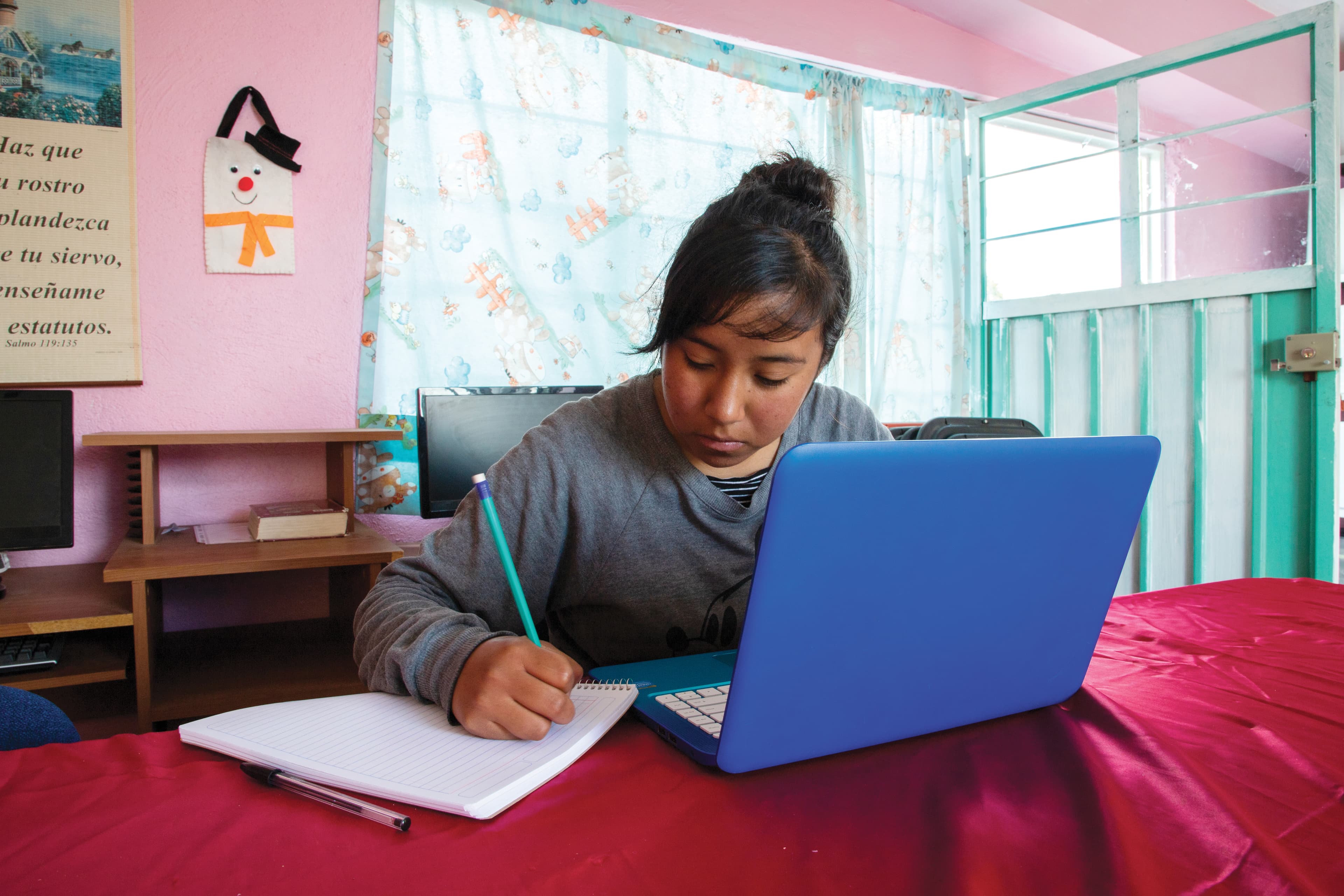 A young woman sits at a table covered in a red tablecloth, working on a blue laptop.