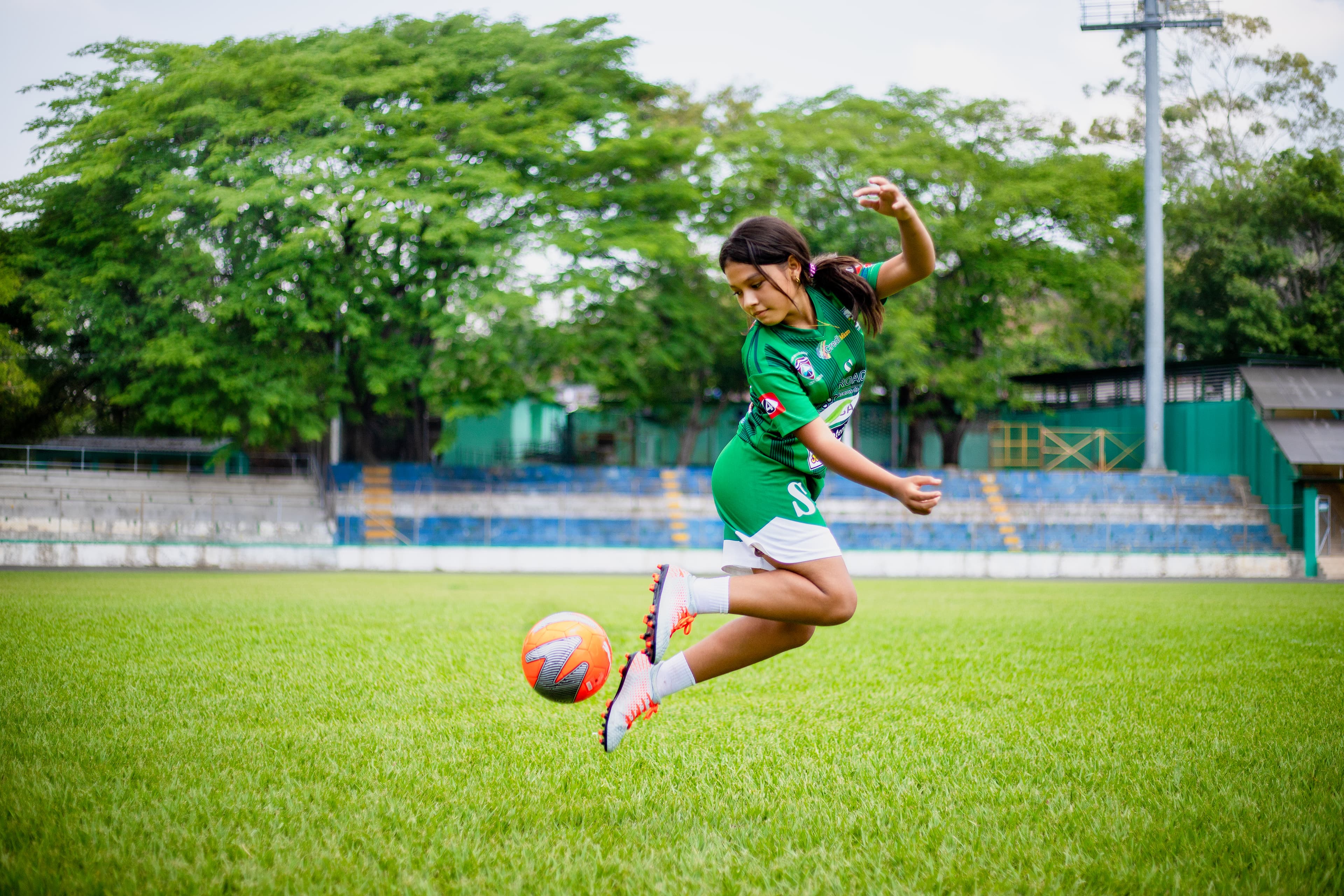 A young girl wearing a soccer uniform jumps in the air to kick a soccer ball.