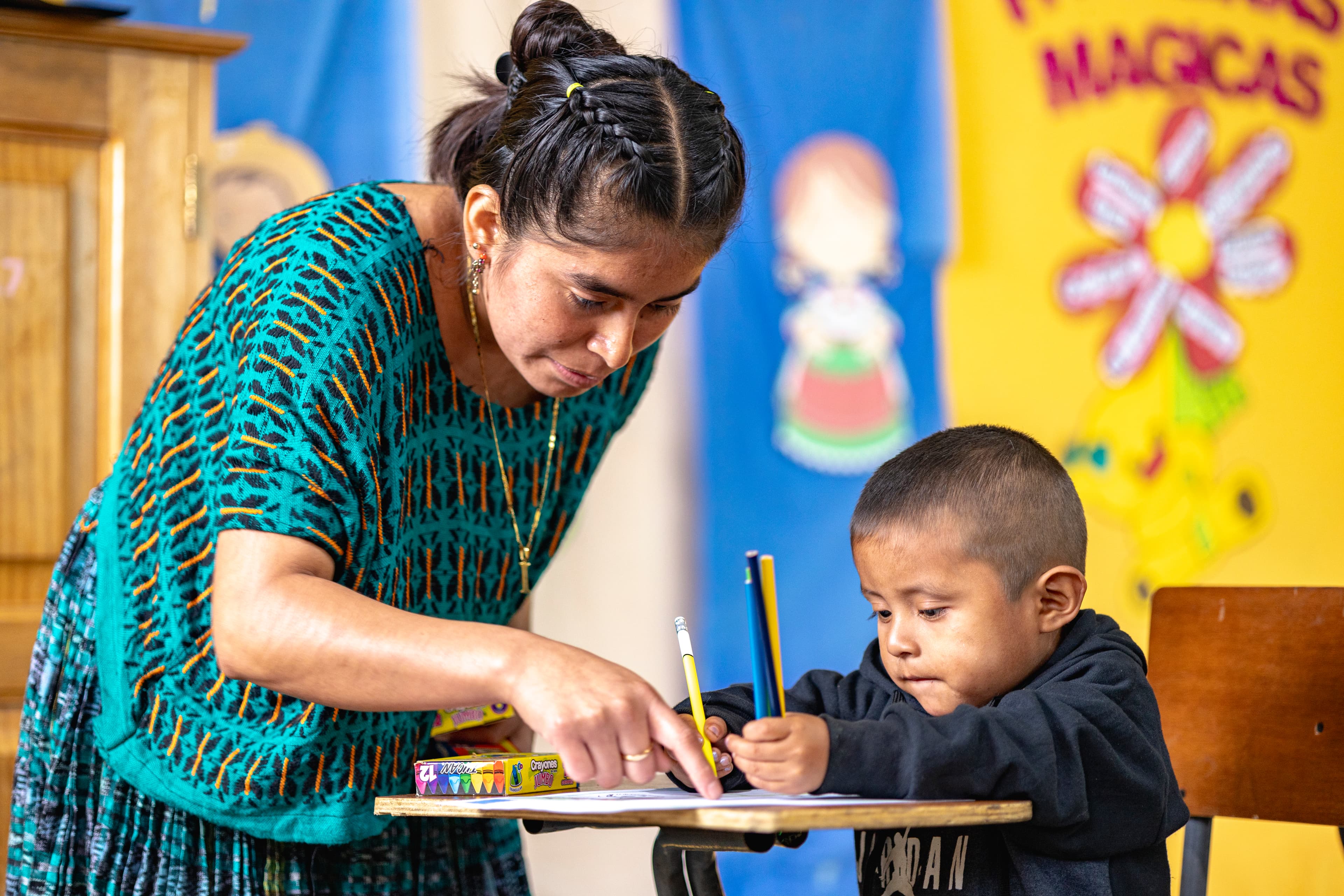 A teacher stands over a little boy as he sits at a wooden desk holding a pencil.