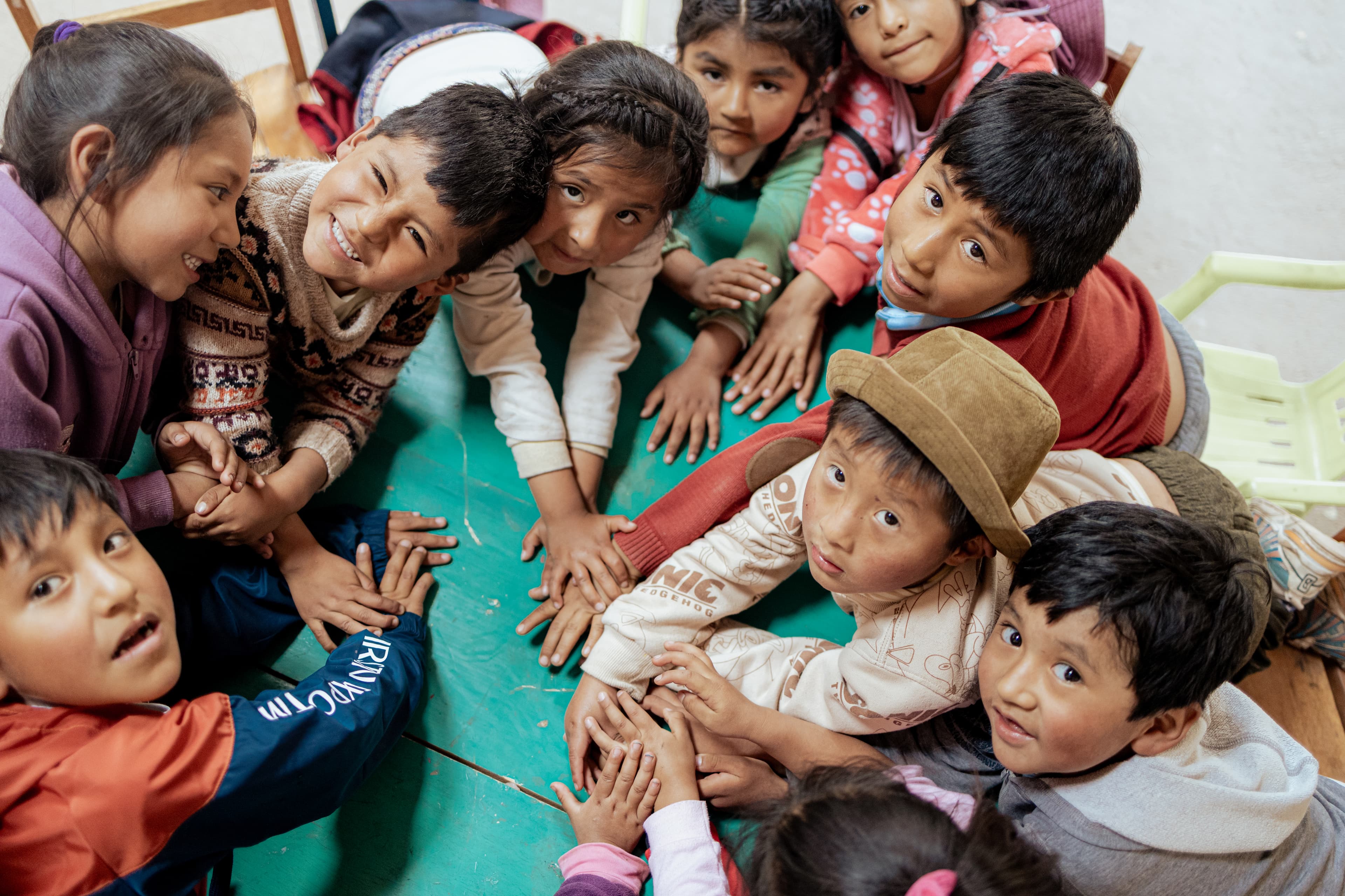 A group of children are circled around and leaning in on a green table looking up at the camera.