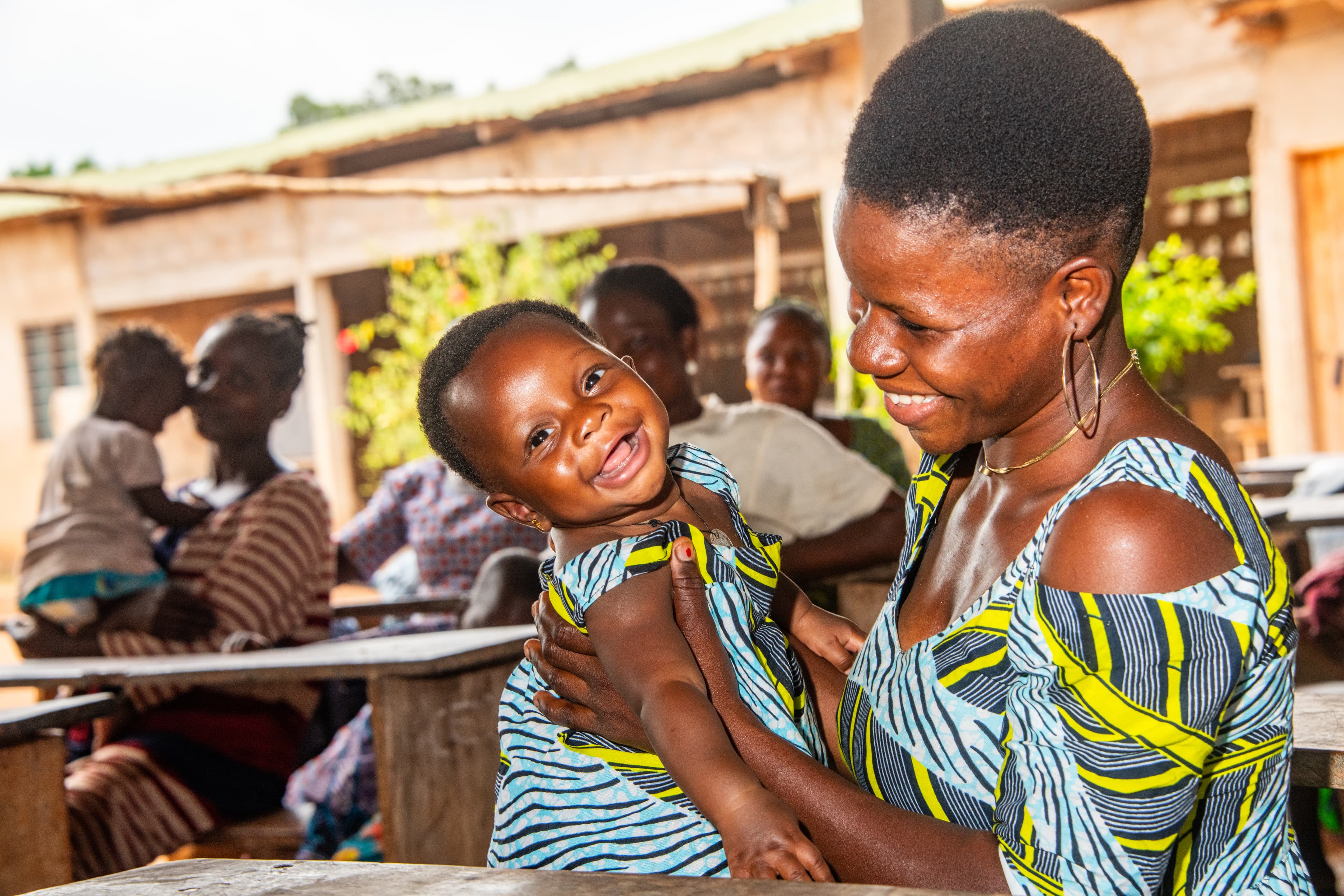 A mother is sitting at a wooden desk holding a baby on her lap and looking at her. They are both smiling. The mother and daughter are wearing matching dresses with a gray, yellow and blue pattern. There are other women and children in the background.