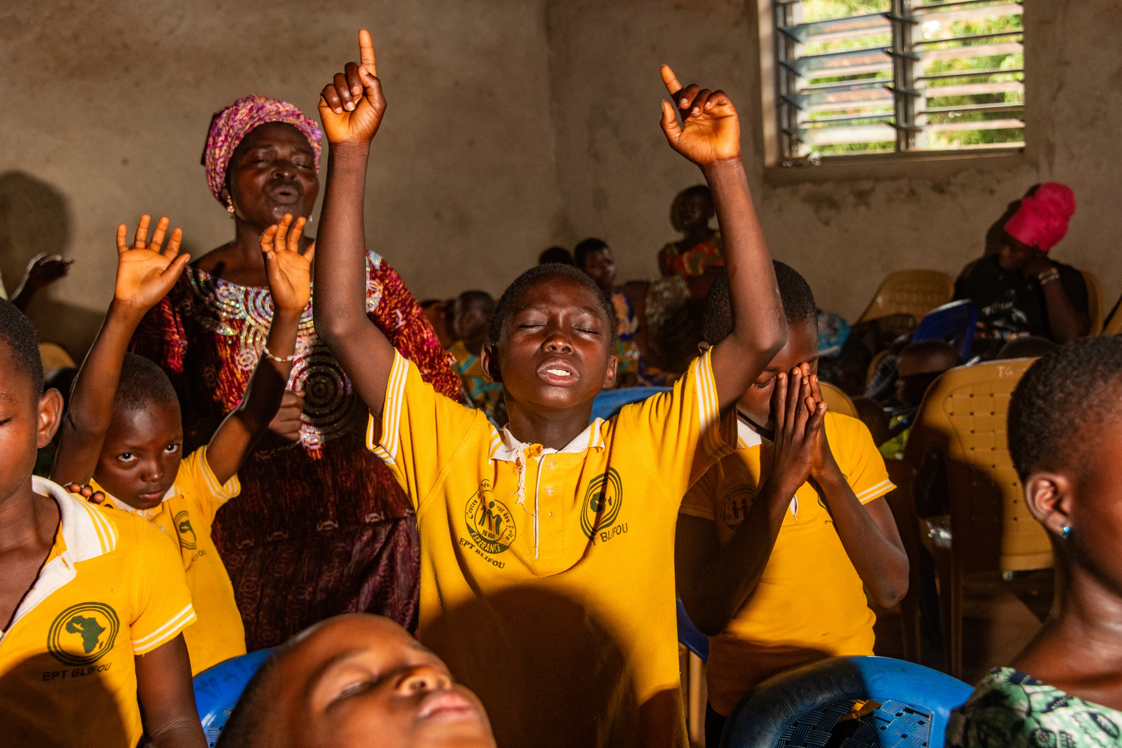 A teen African boy surrounded by other children lifts his hands in the air with his eyes closed.