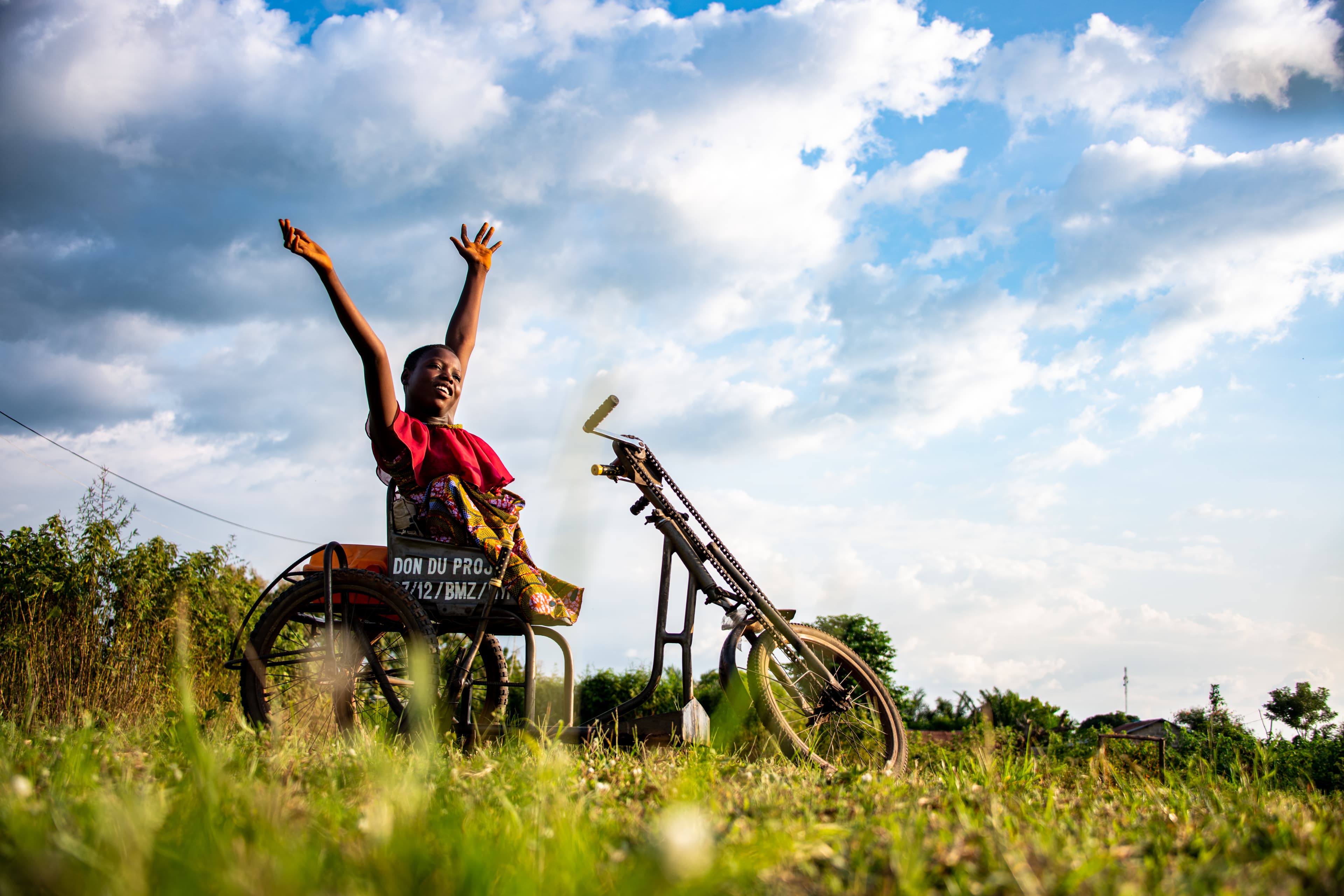 An African teen girl sits in a wheelchair with her hands in the air as she smiles.