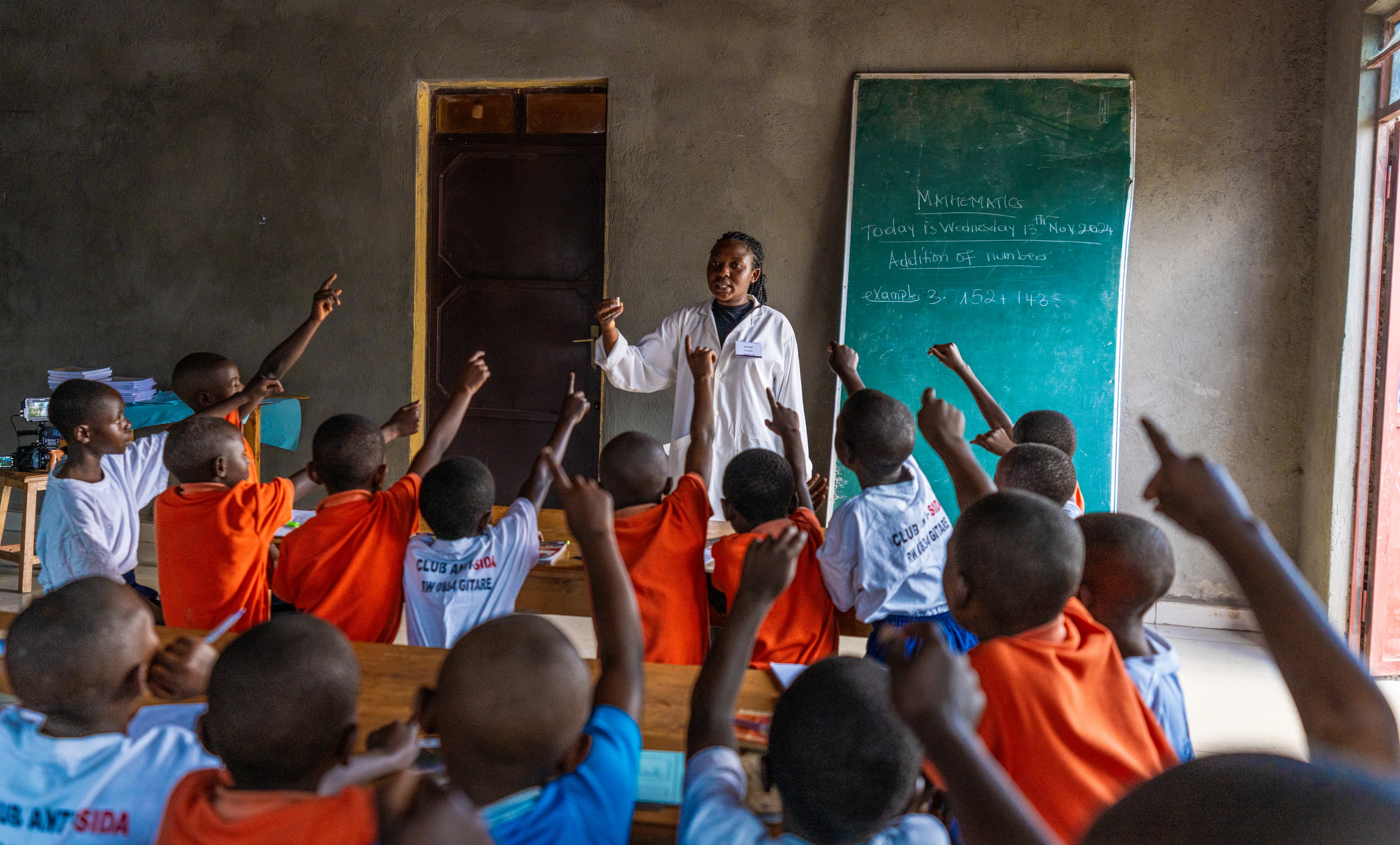 A Compassion alumni is standing at the front of a classroom facing children who are sitting on stools with their hands raised.
