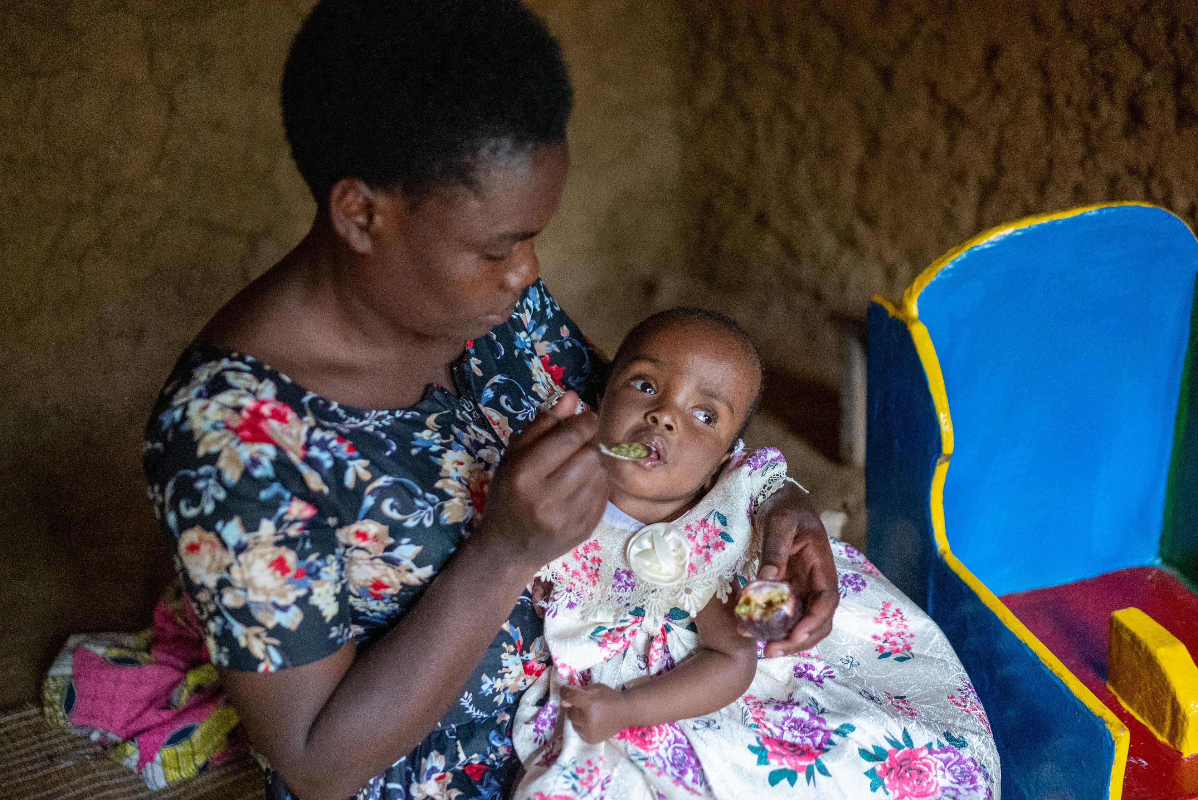 Mother is sitting on a chair holding her daughter and feeding her with a spoon.