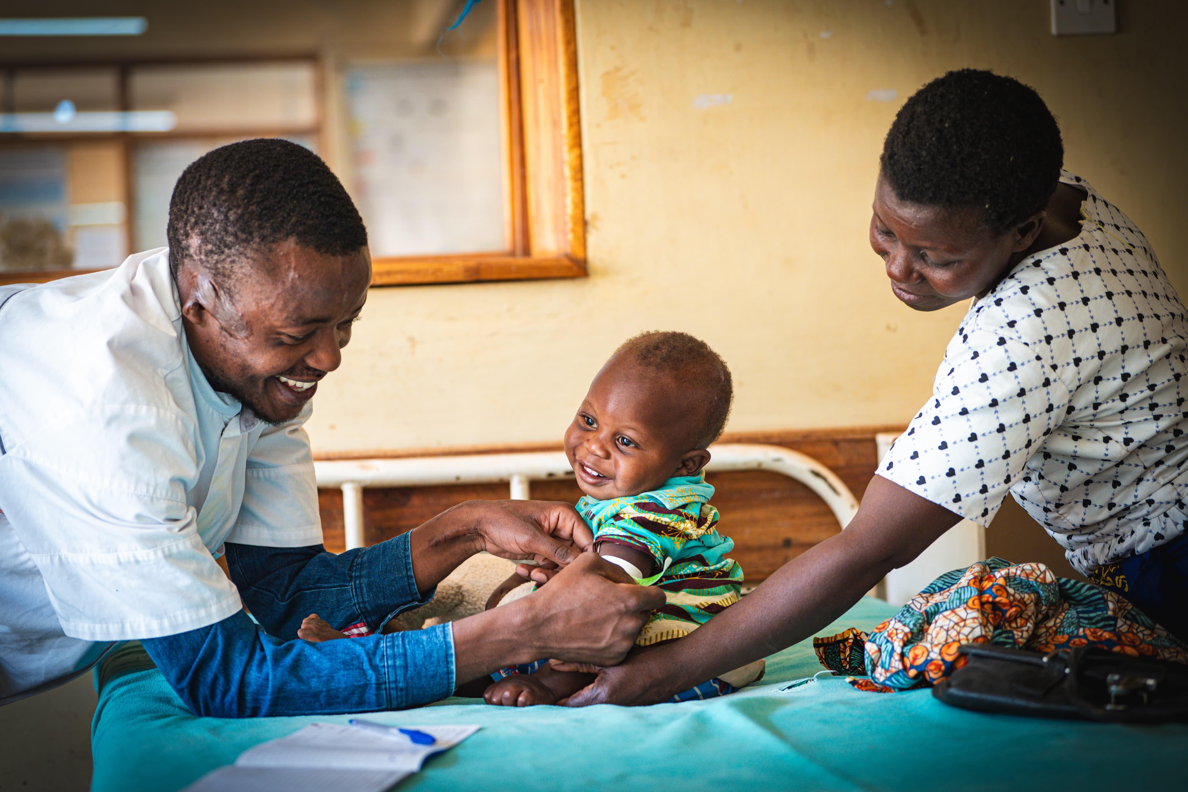 A baby is sitting on a hospital bed receiving care from a doctor while his mother comforts him.