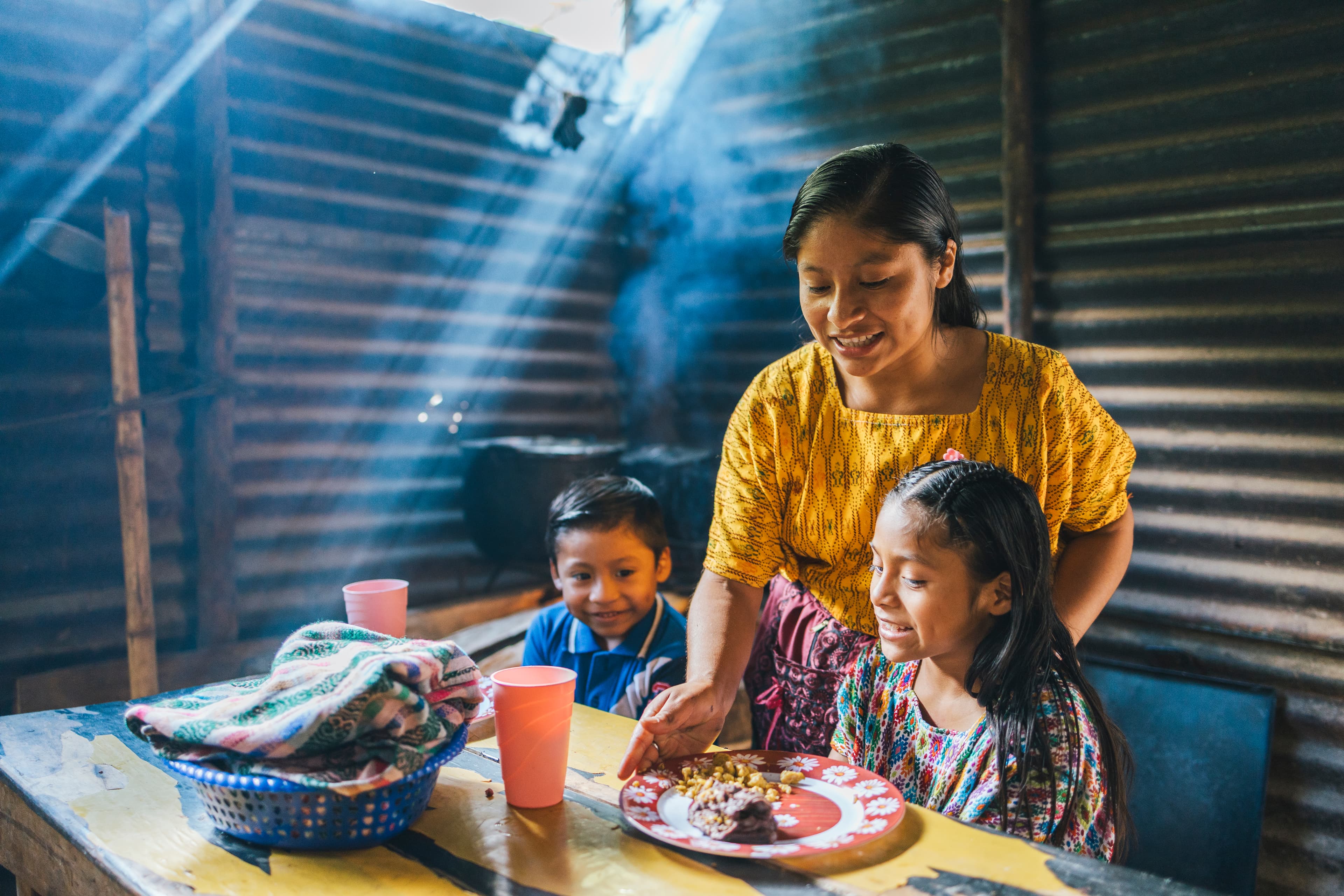 A girl in a colorful patterned shirt and a boy in a blue polo shirt are preparing to eat the breakfast their older sister is serving to them at a table. Light is streaming in through their home, which has tin walls.