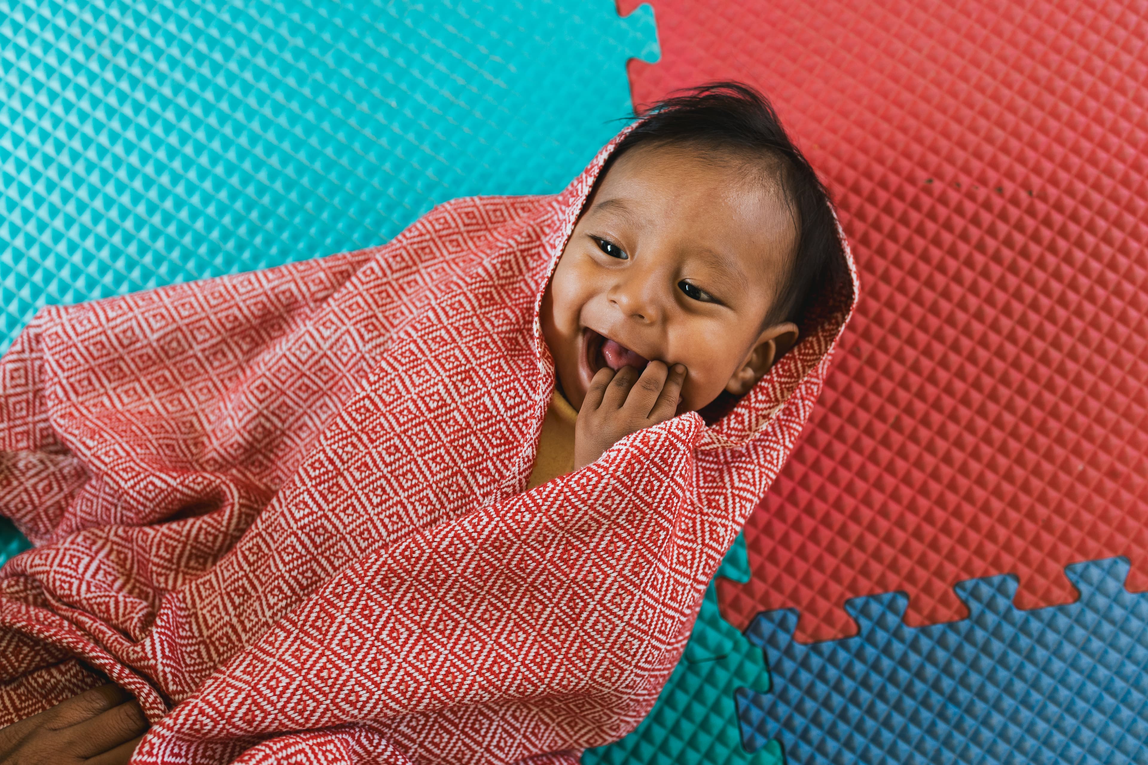 A baby boy wrapped in a red, woven cloth lies on colorful foam panels.