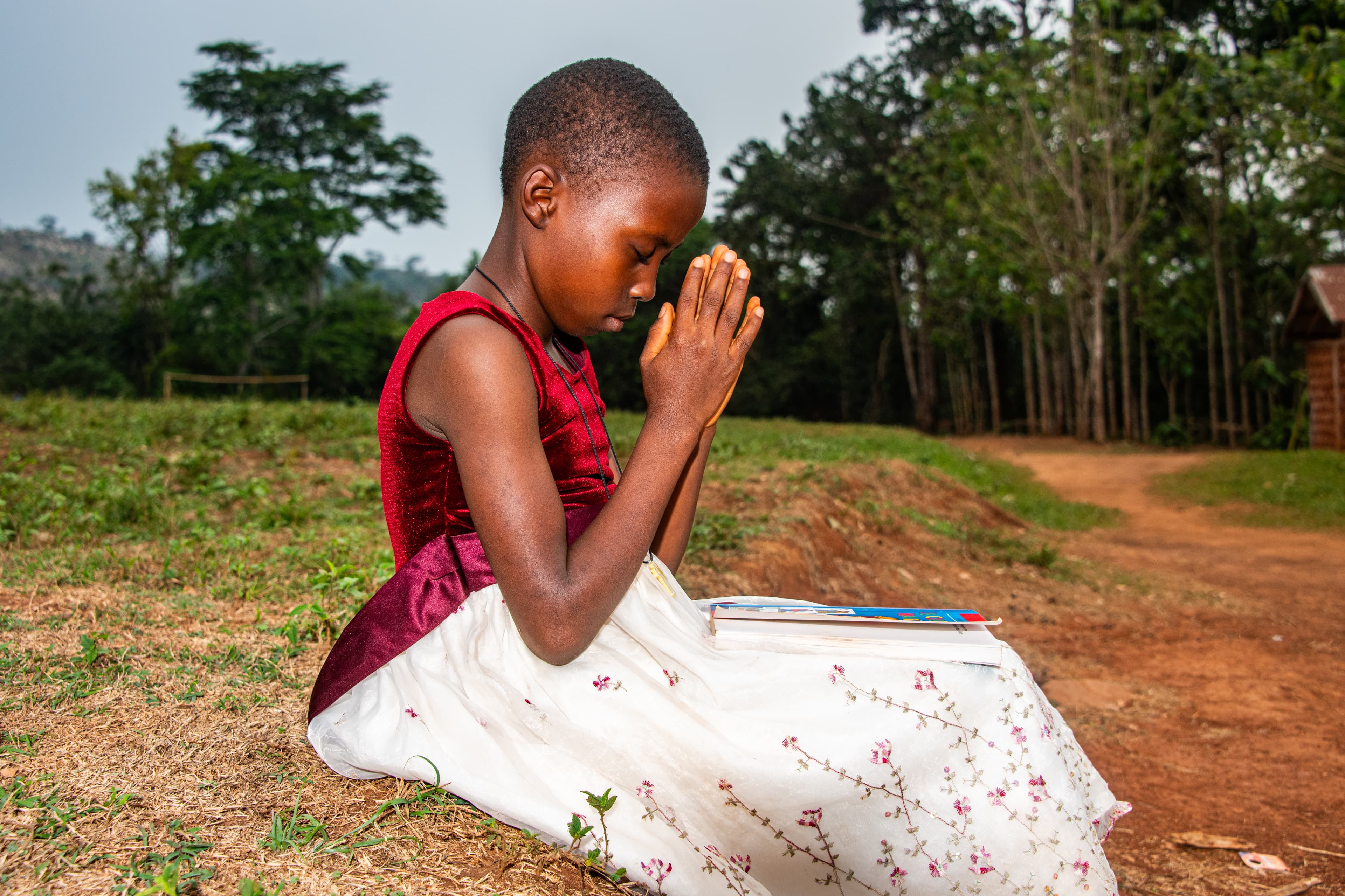 A young African girl wearing a floral dress prays in a field with a Bible on her lap.