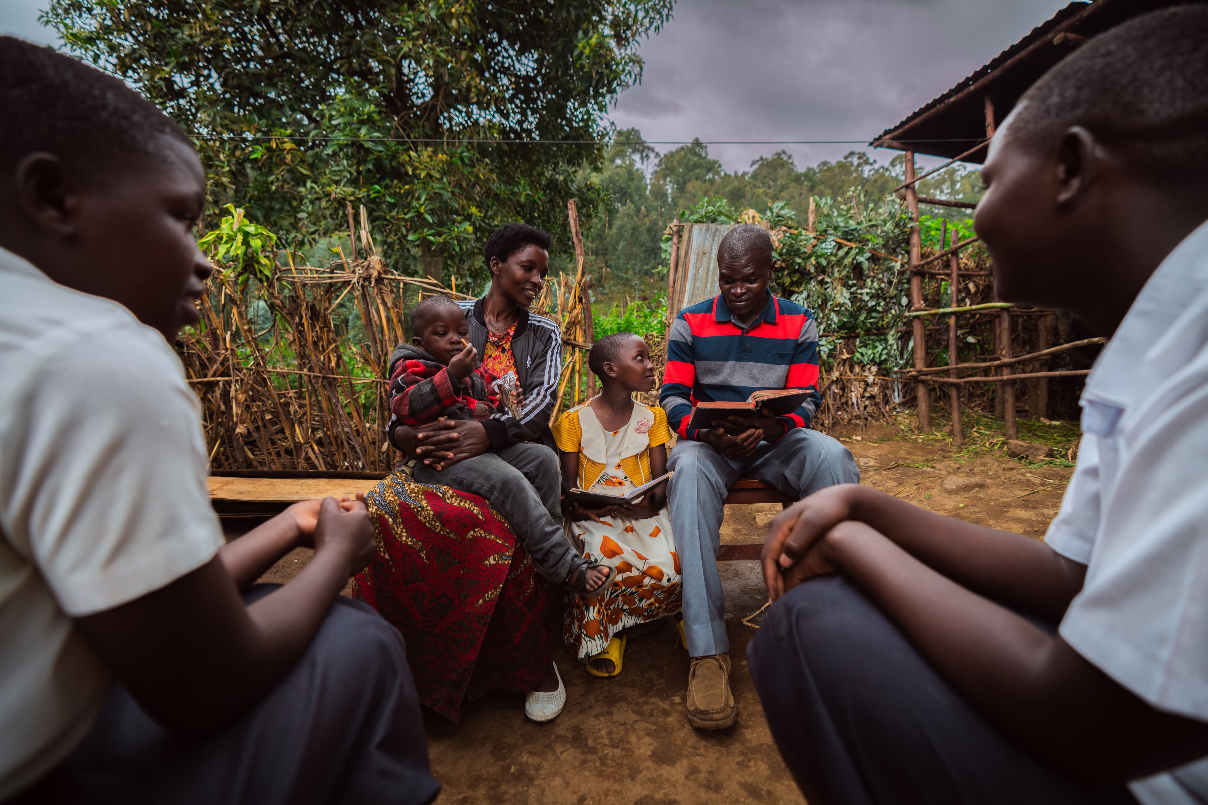A man, woman, girl and toddler boy sit outdoors on a wooden bench reading Bibles. Two older boys sit opposite of them in the foreground.
