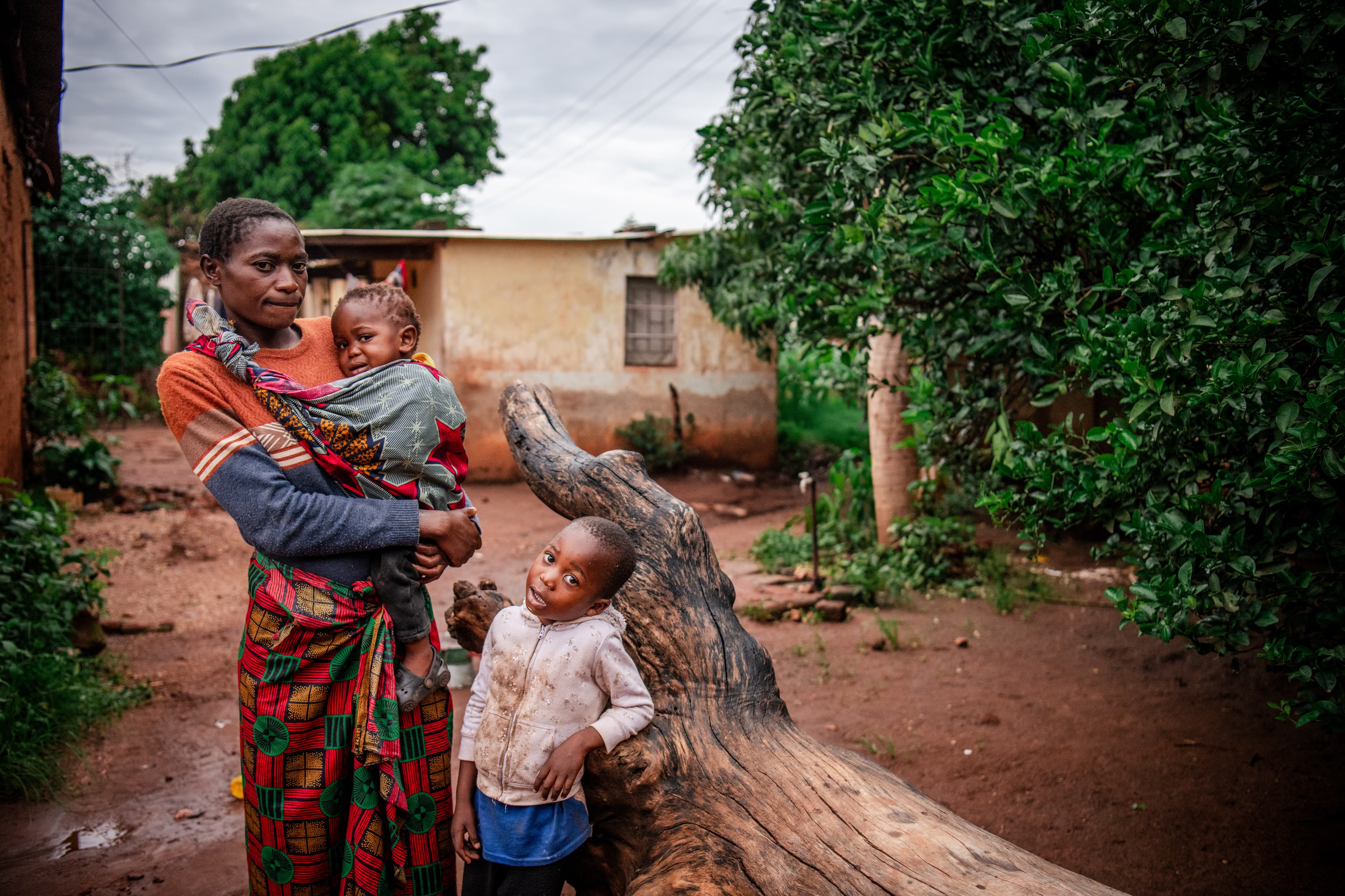 Beauty and her children are standing outside their house. Beauty is wearing her daughter Alice in a colorful sling, while her other child leans against a log. They are looking at the camera.