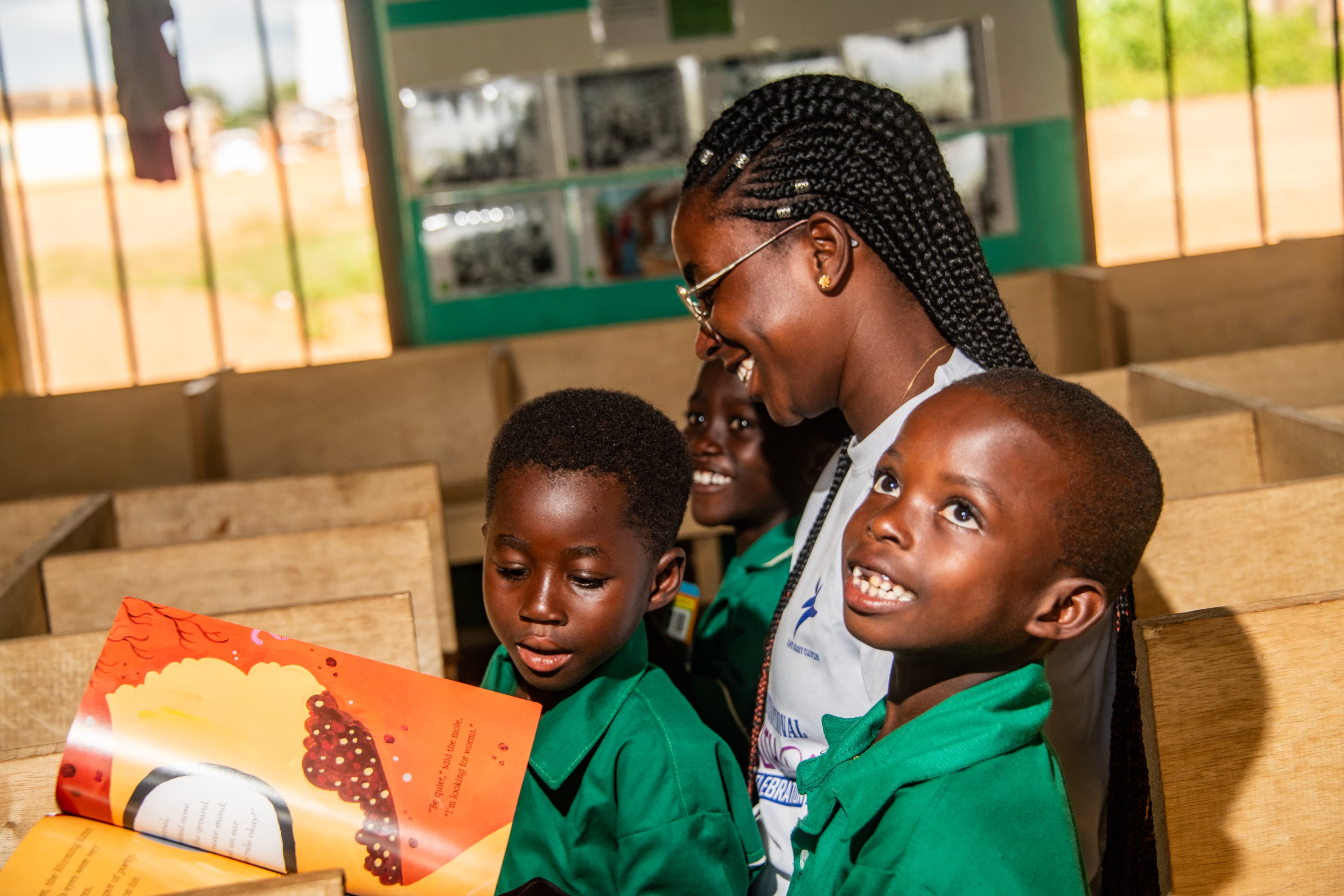 Three children wearing green shirts read a picture book with a woman wearing a white shirt, sitting at wooden desks.