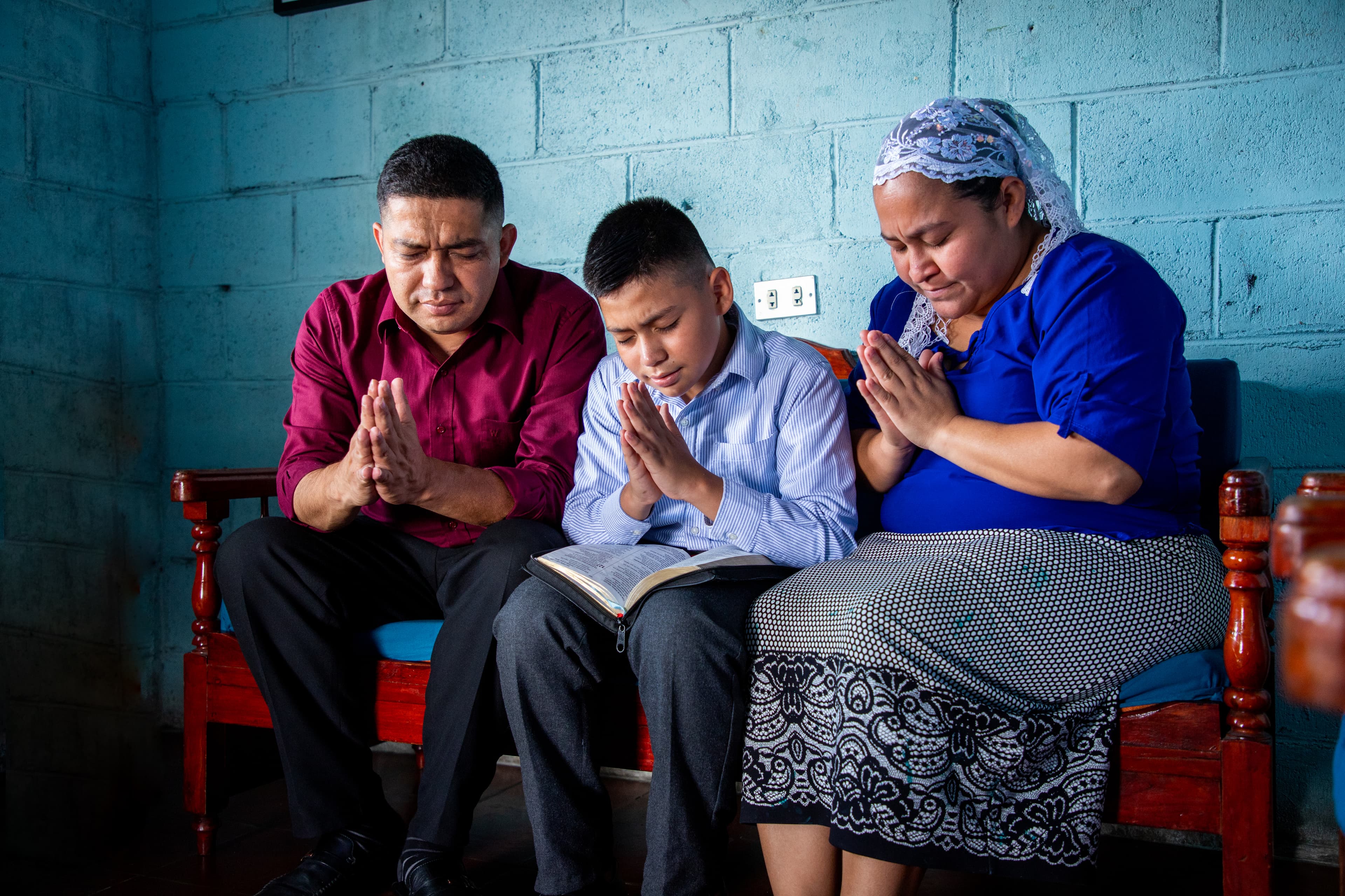 A mother, father and son are praying together on a wooden bench. The son is wearing a blue and white striped shirt. He is sitting between his mother, wearing a blue shirt, and his father, wearing a maroon shirt. The wall behind them is blue.