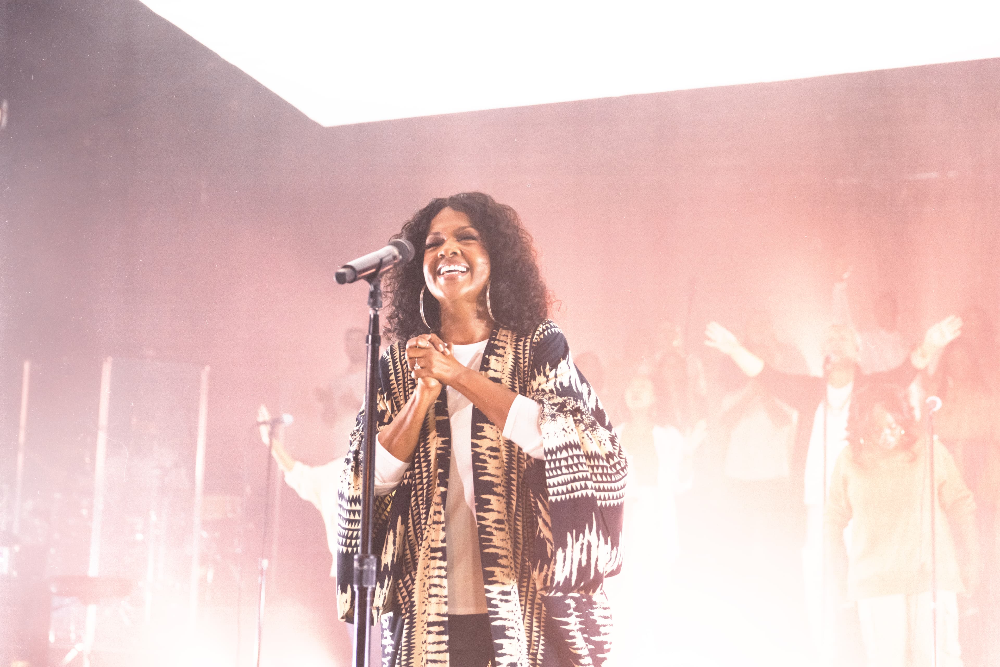 CeCe Winans is smiling as she stands behind a microphone on a brightly lit stage.